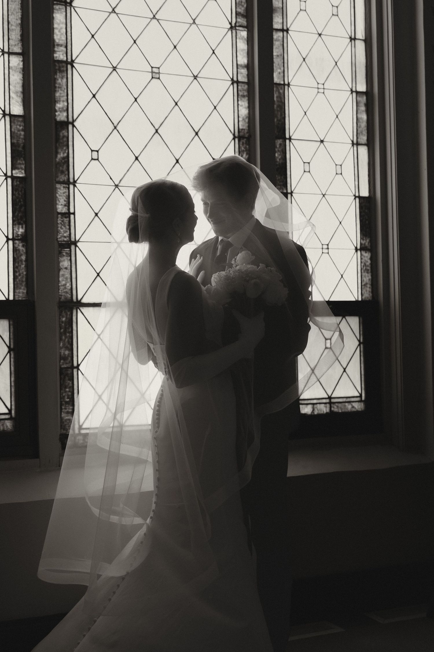 Black and white photo of bride and groom silhouetted against stained glass church window on wedding day.