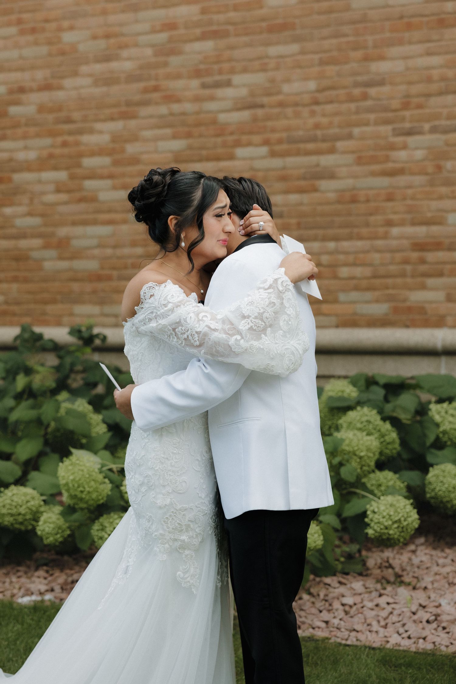 A couple embraces in their wedding attire against a brick wall with green hydrangea bushes.