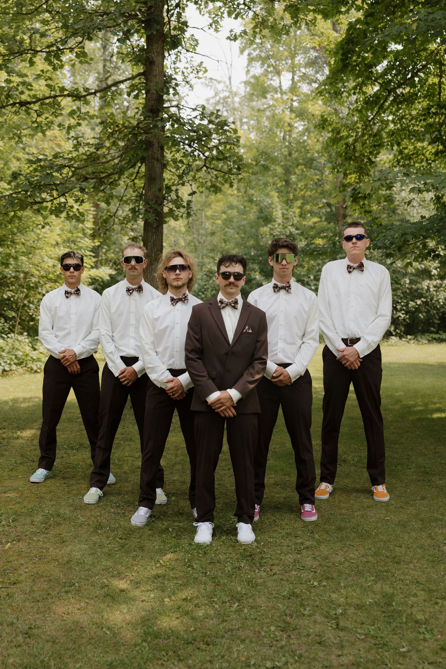 Groomsmen in black and white attire posing casually on grass under trees.