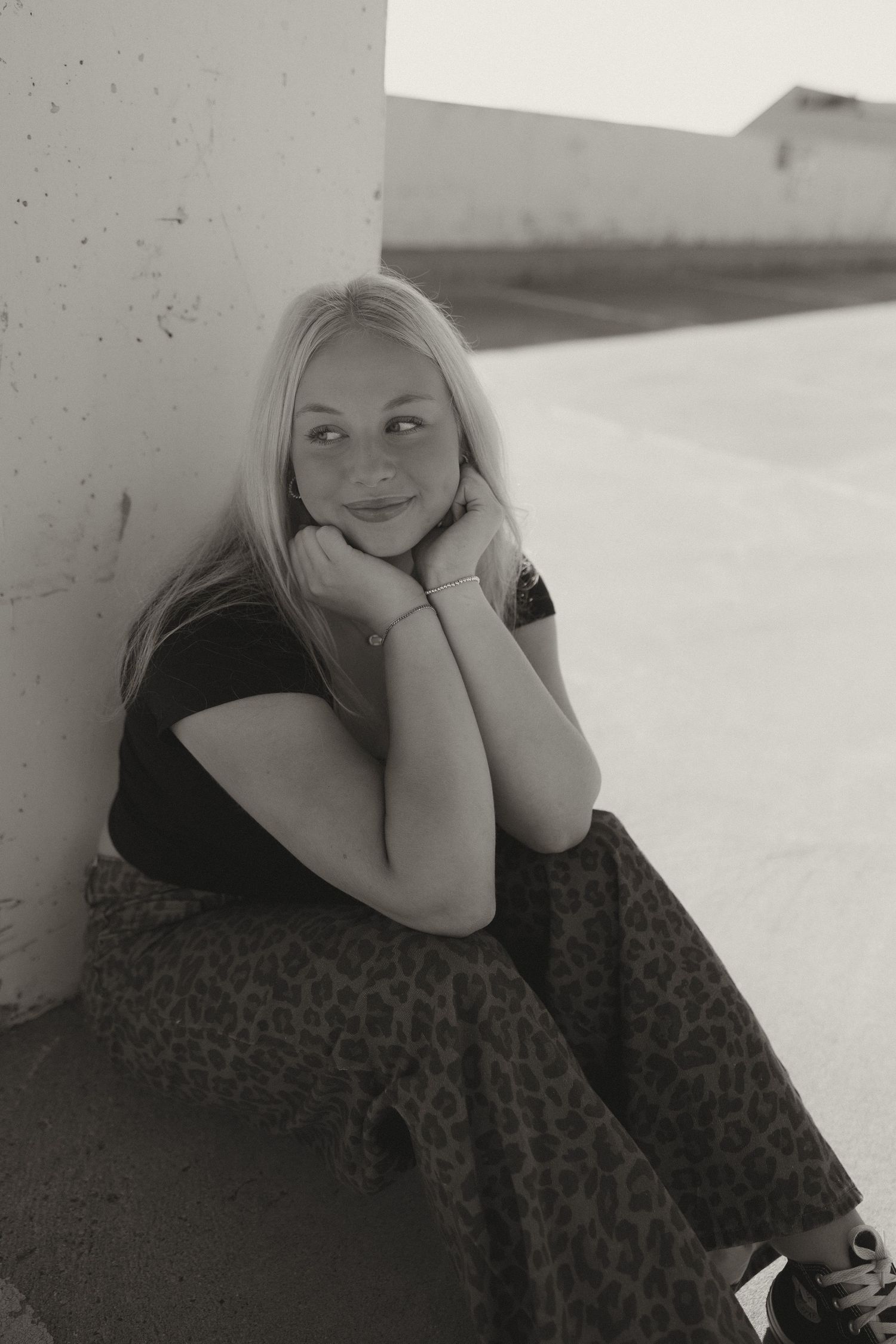 Black and white portrait of a blonde teen resting chin on hands while sitting against a wall outdoors.