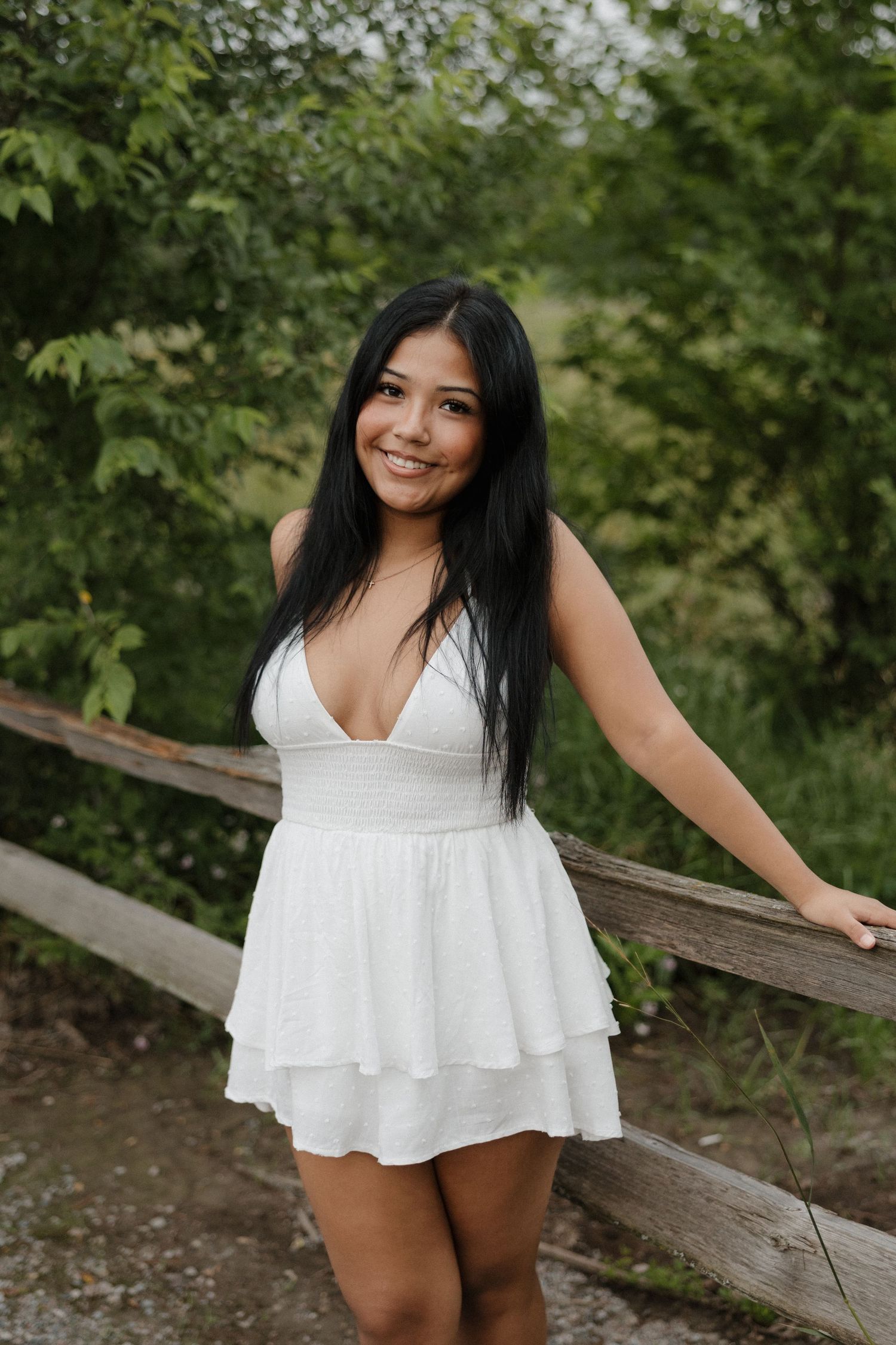Smiling young woman with black hair in white mini dress leaning on rustic wooden fence outdoors.