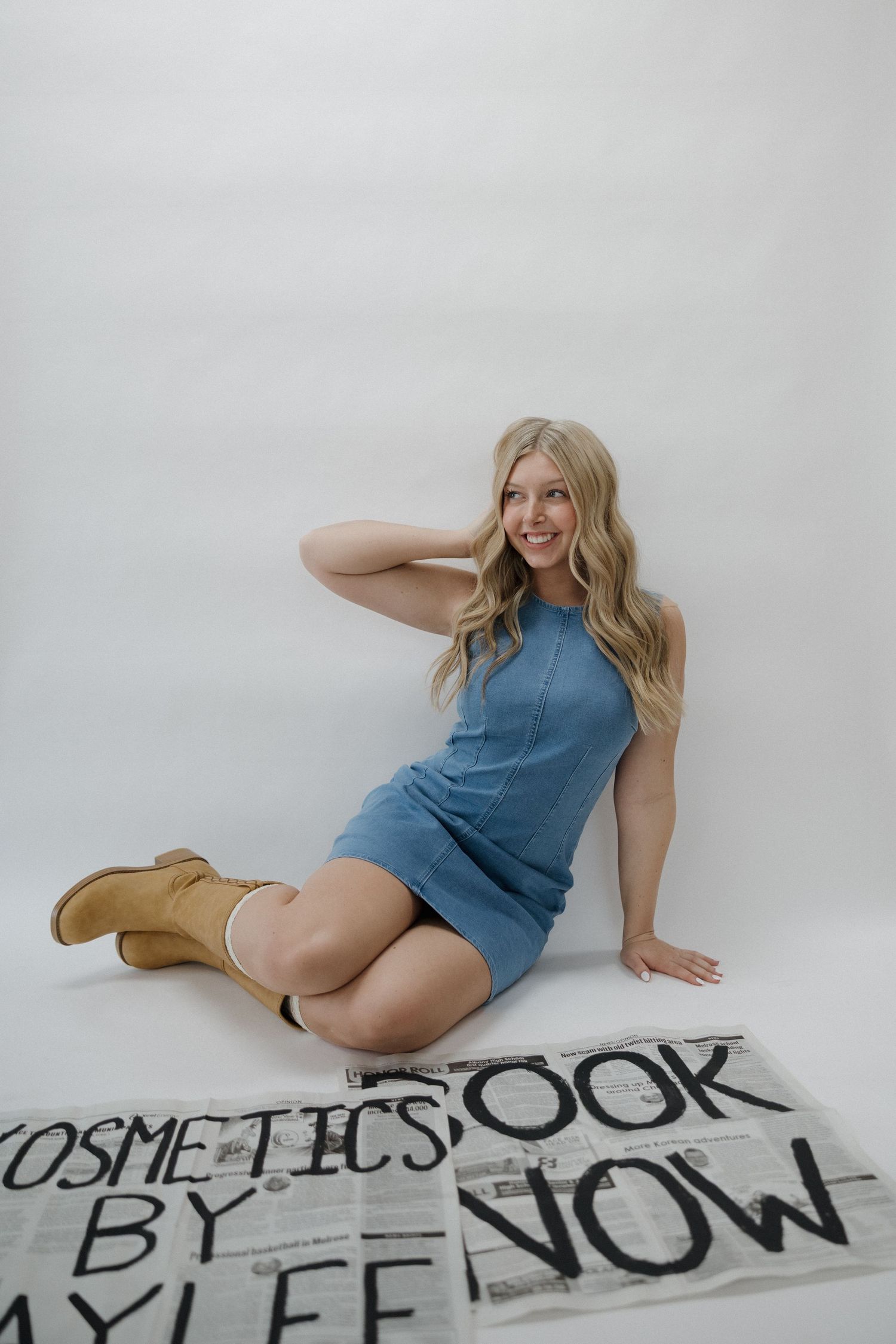 Smiling blonde woman in blue dress and tan boots sitting on floor beside cosmetics brand newspaper signs.