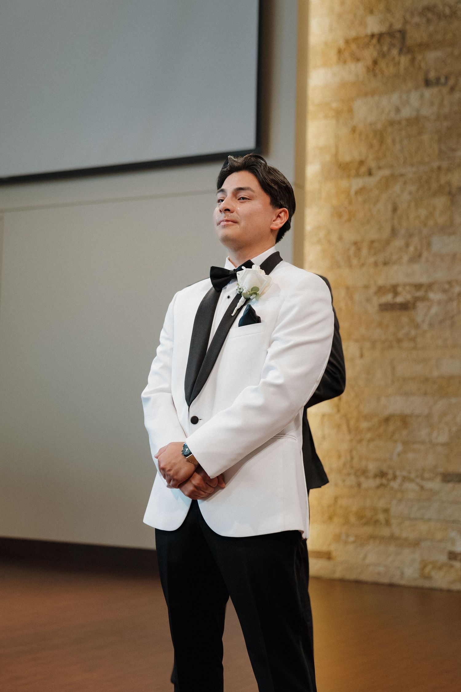 Formal portrait of someone in a white dinner jacket with black bow tie against a stone wall.