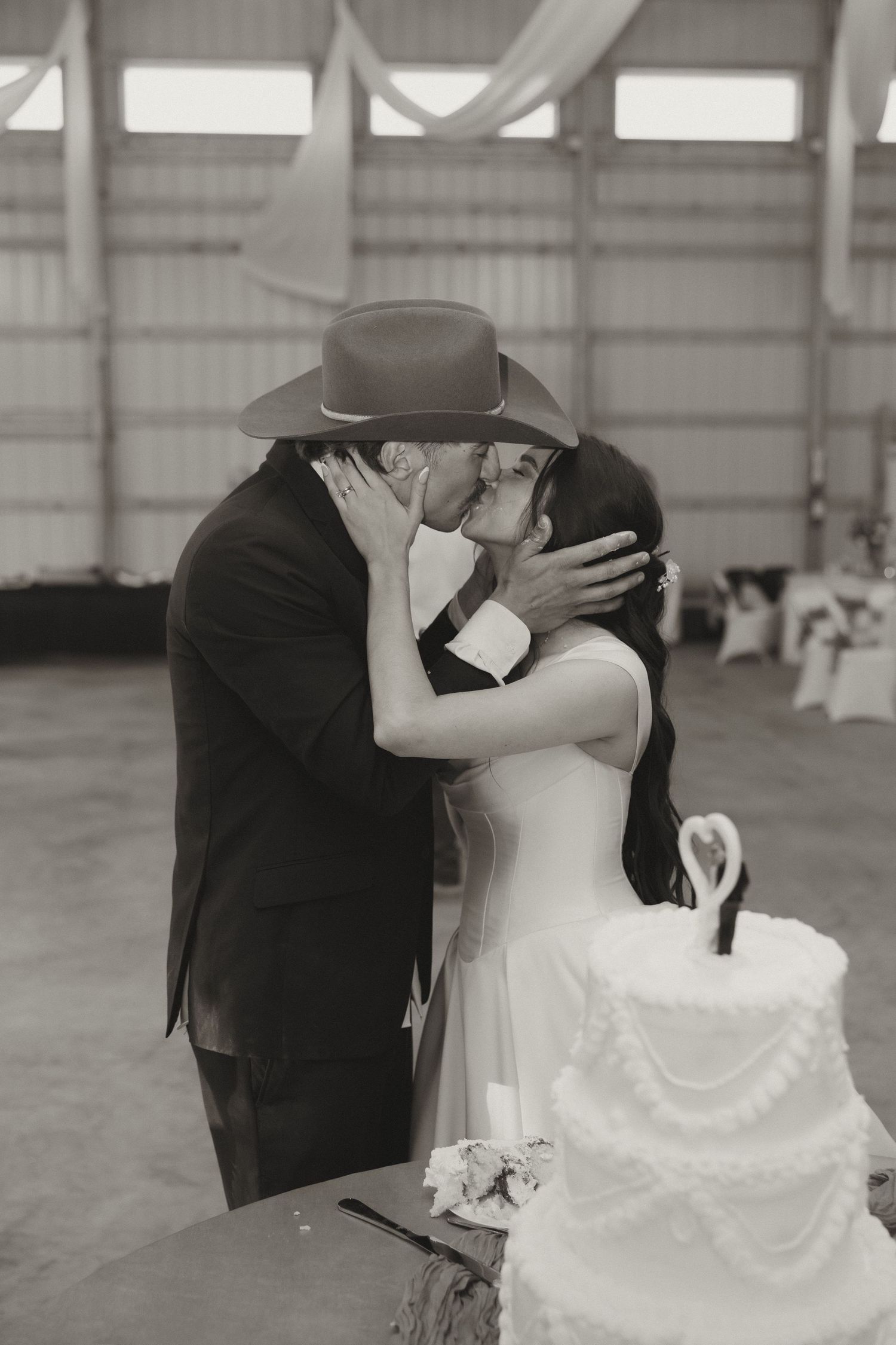 A romantic black and white wedding photo showing a tender moment beside an elegant white tiered cake.