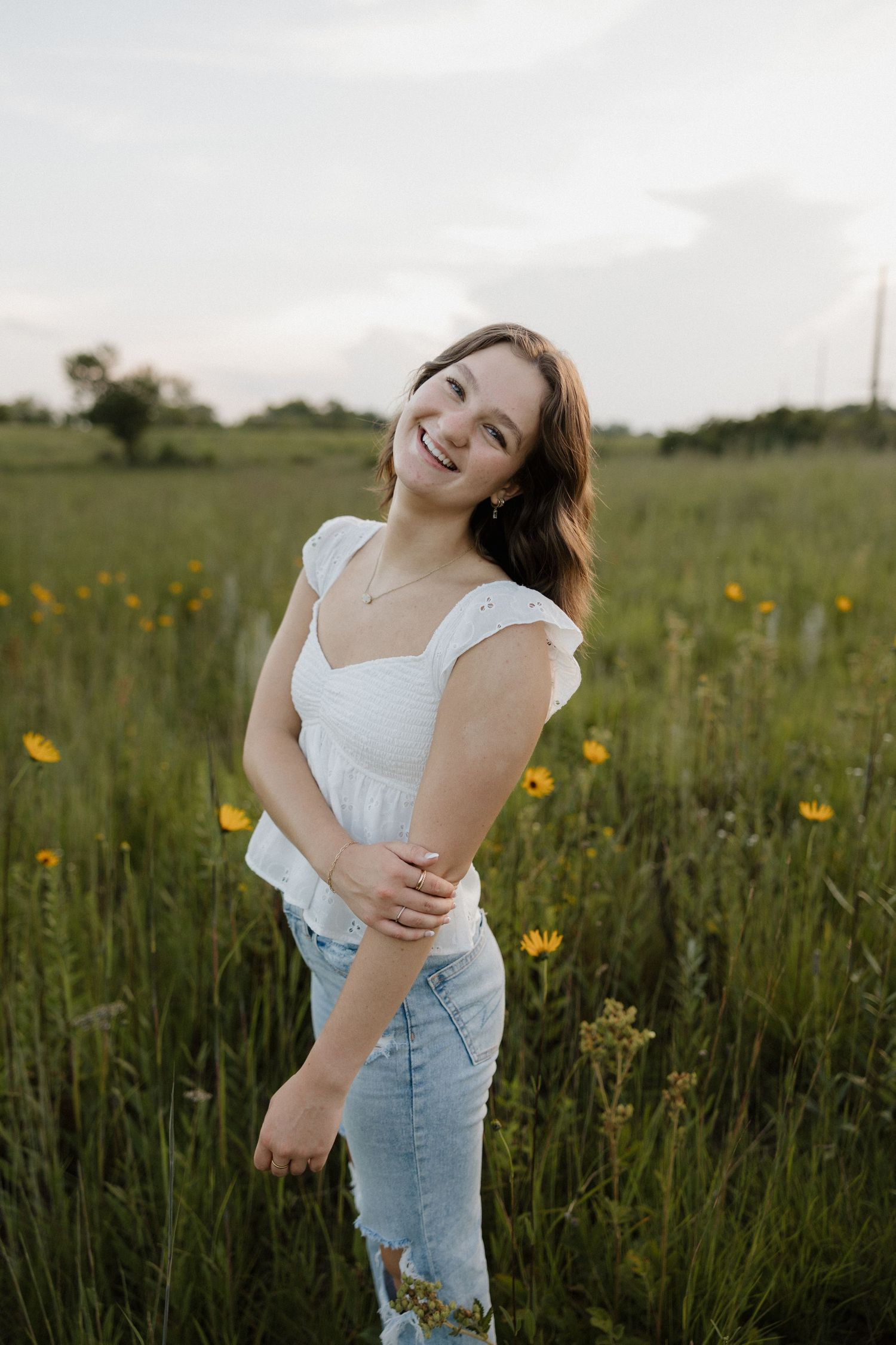 Smiling young woman in a white top and ripped jeans posing in a green wildflower field.