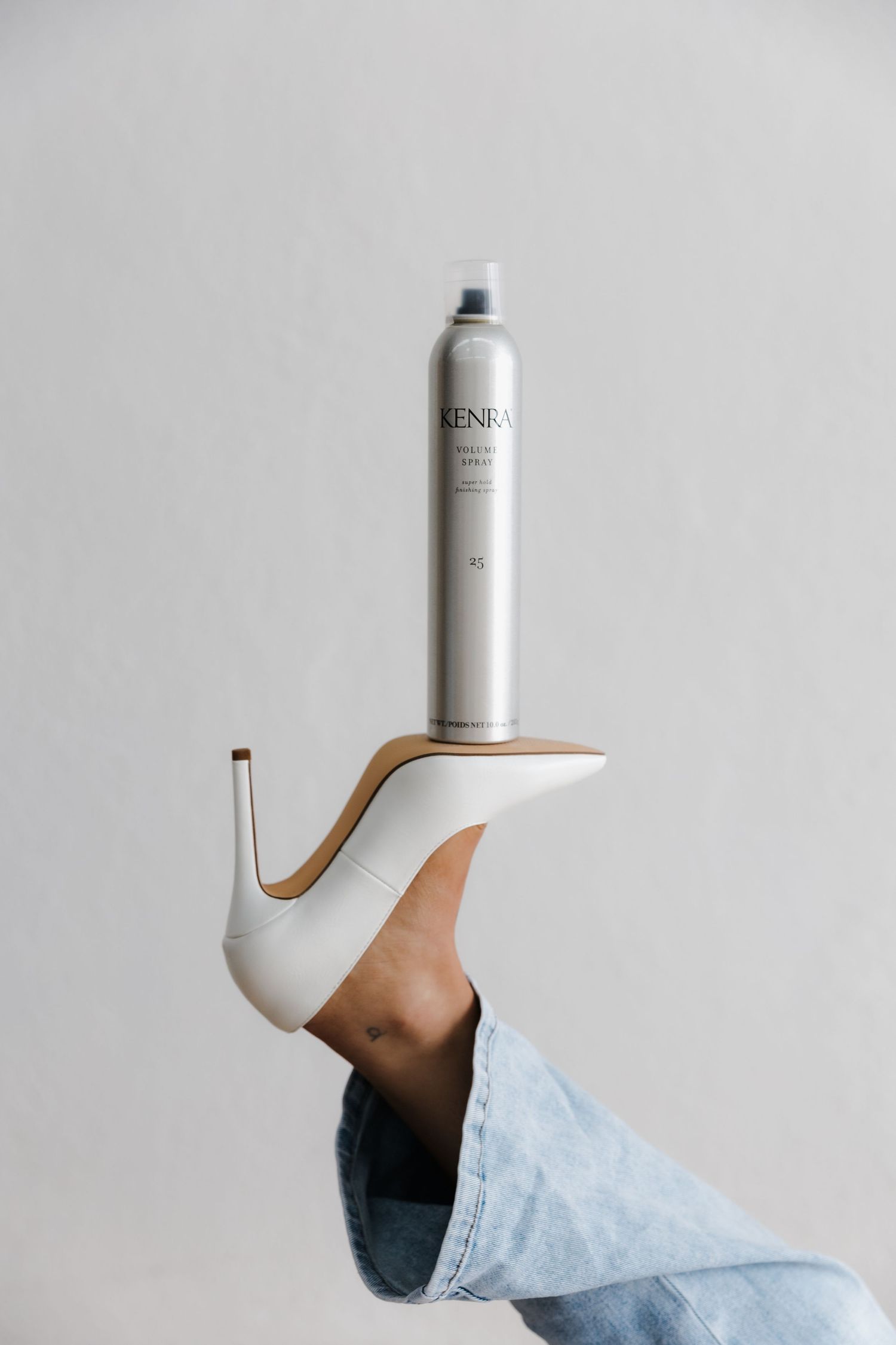 Leg in white stiletto heel balancing a Kenra hair spray can against a gray background.