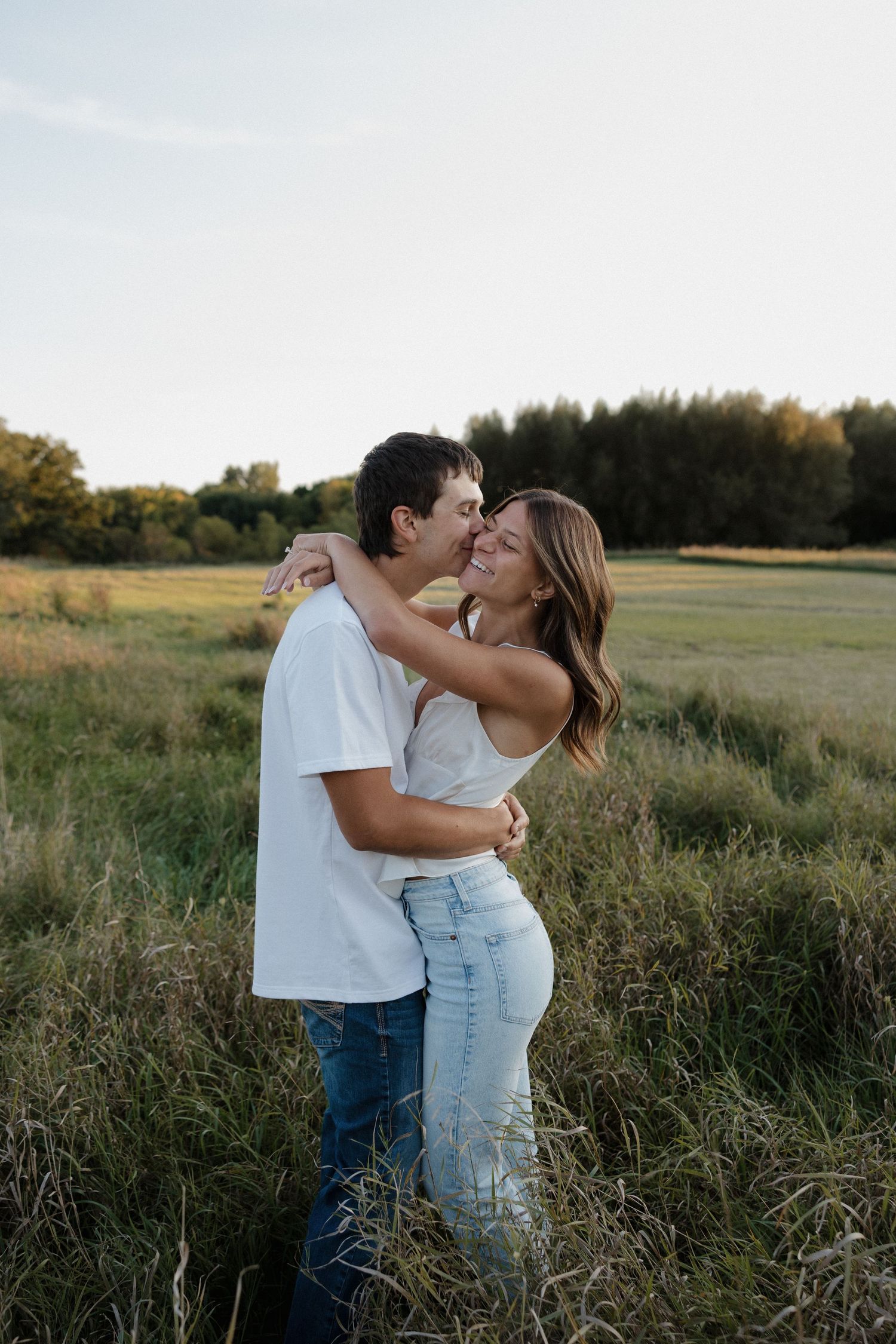 Joyful couple hugging and laughing together in a golden hour green field during engagement photos.