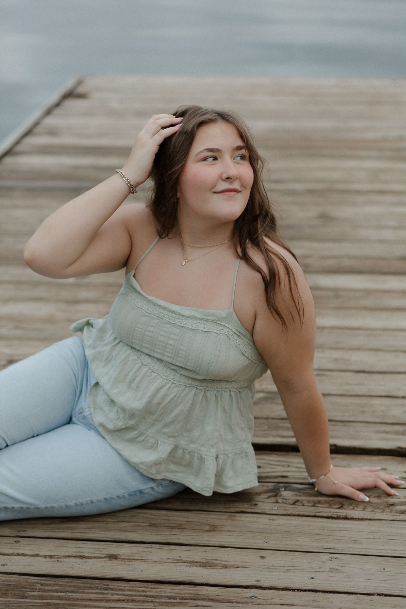 Young woman in sage green top and jeans posing on wooden dock by calm water.