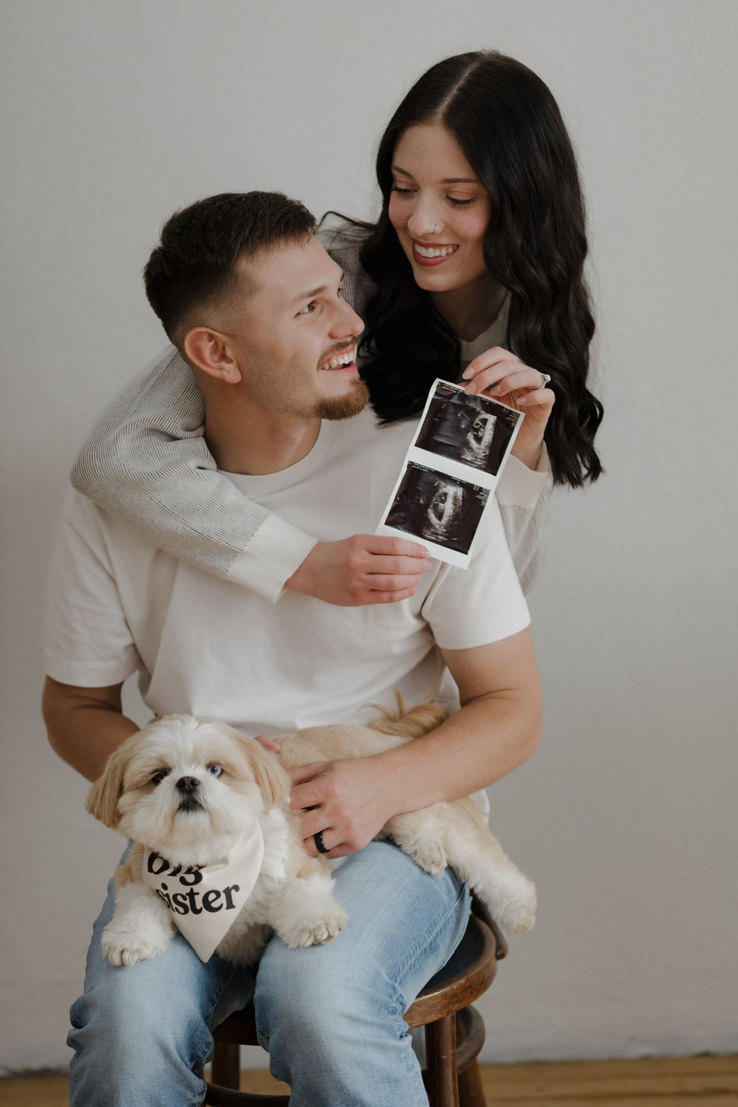 Smiling couple holding ultrasound photos with Shih Tzu dog wearing big sister bandana.
