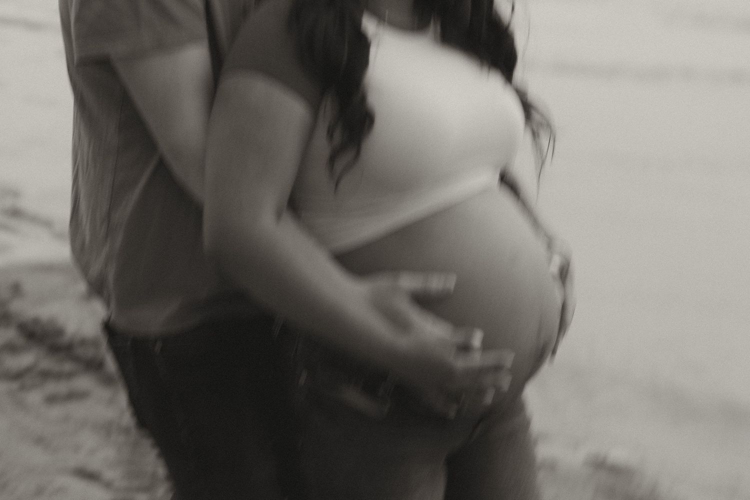 Black and white close-up of couple tenderly holding pregnant belly together outdoors.