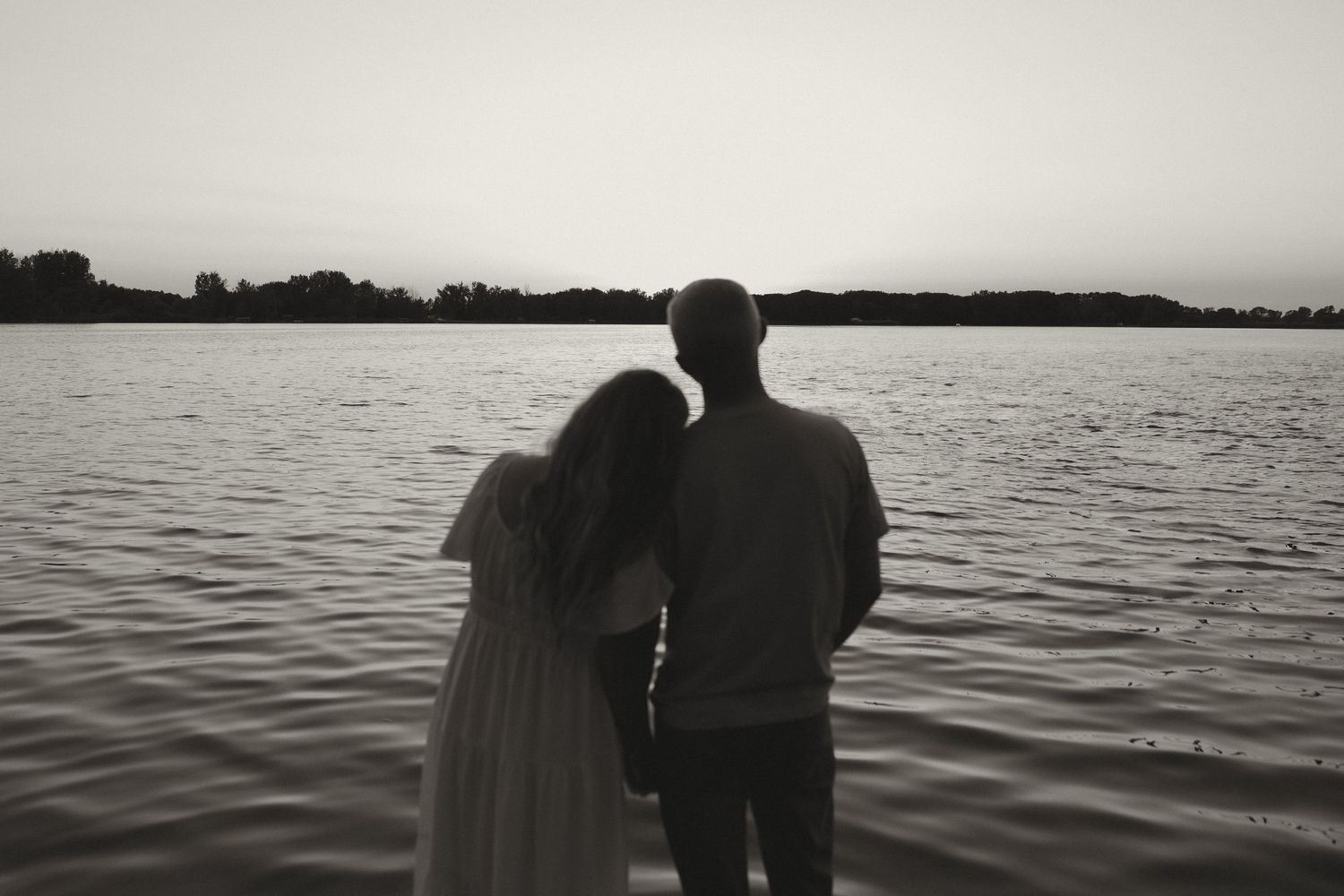 Romantic couple standing together in silhouette, gazing at a serene lake at sunset in black and white.