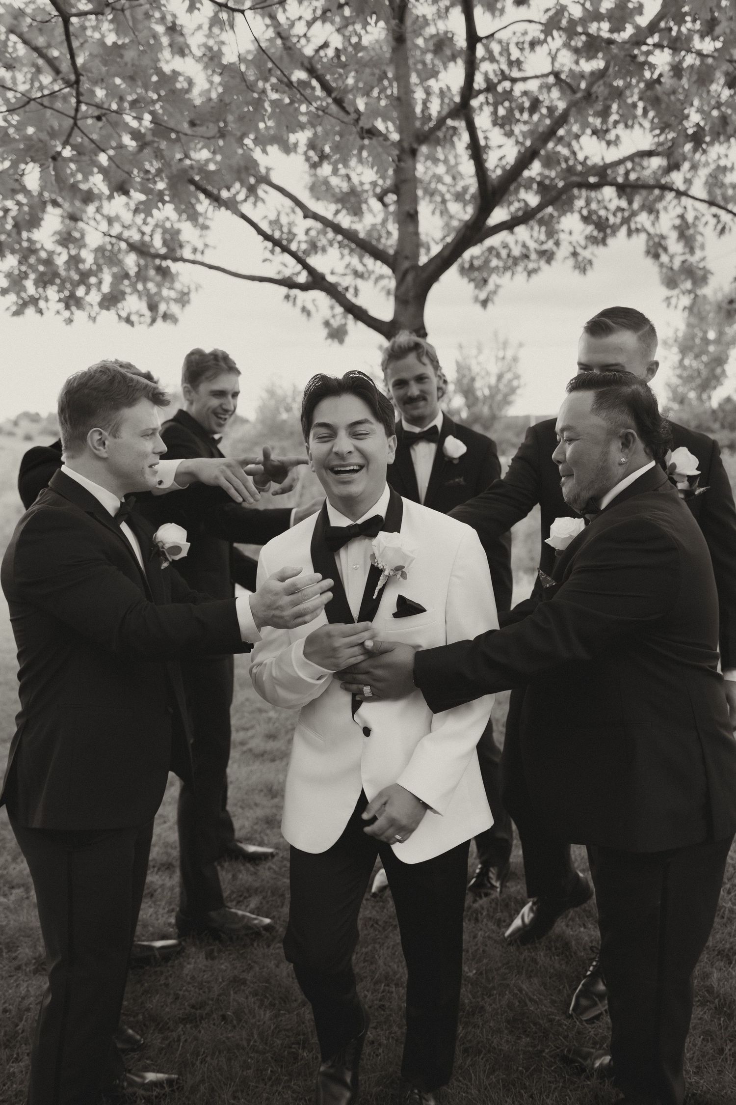 Groomsmen in black suits and white jacket celebrate outdoors under trees.