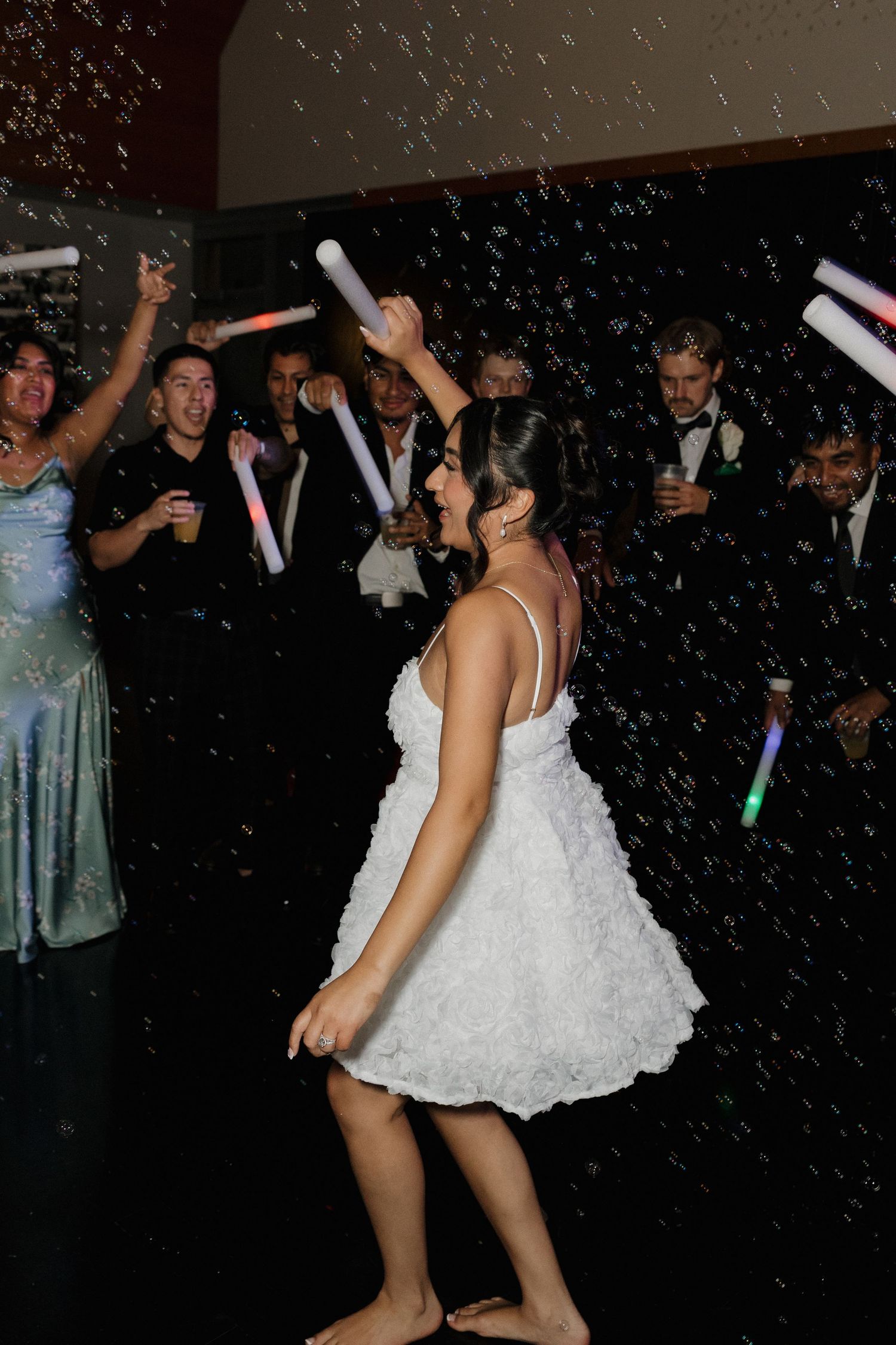 Bride in white dress dancing energetically at wedding reception with glow sticks and party lights.