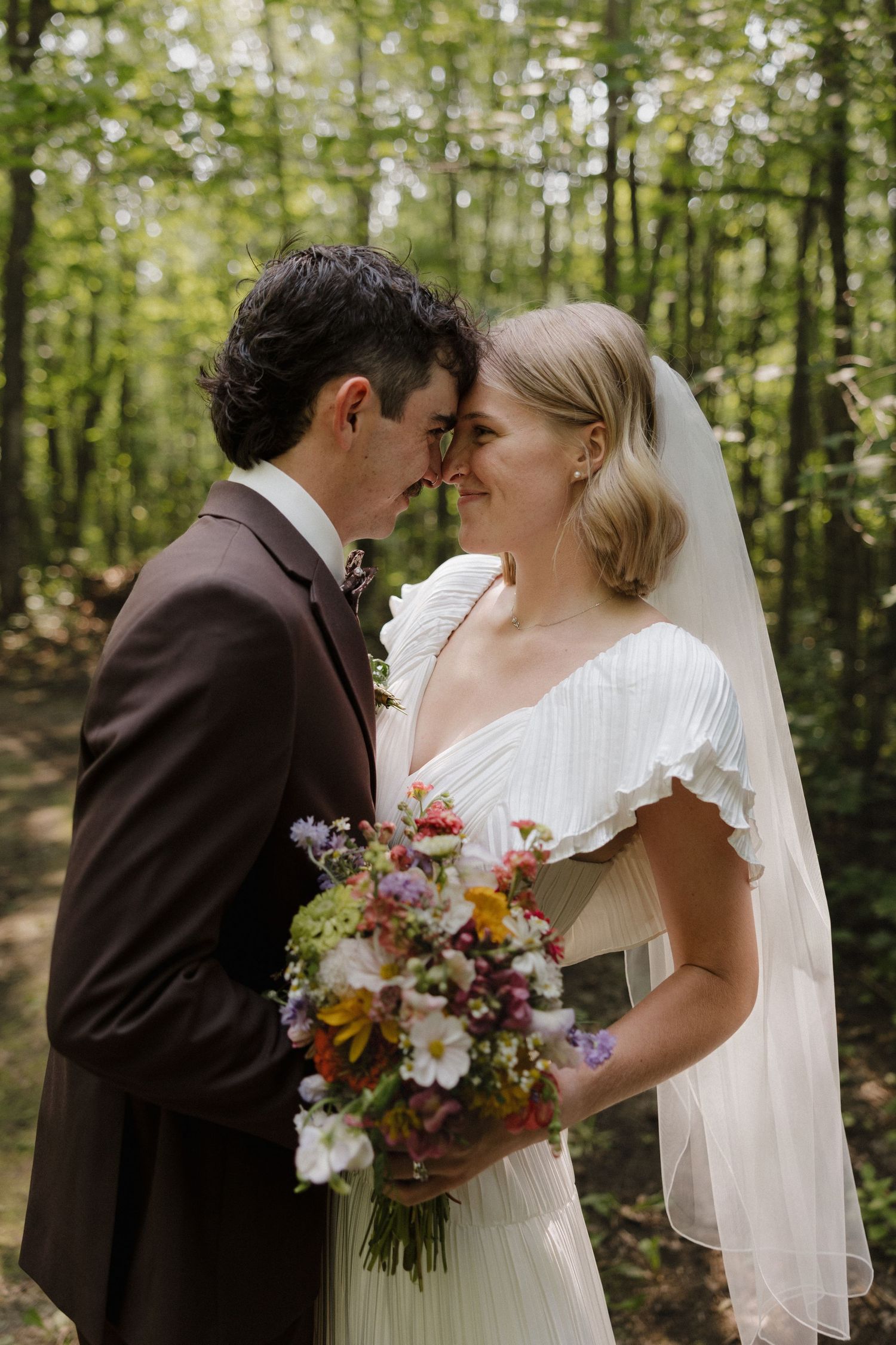 Intimate wedding ceremony moment captured in a sunlit forest with colorful wildflower bouquet.