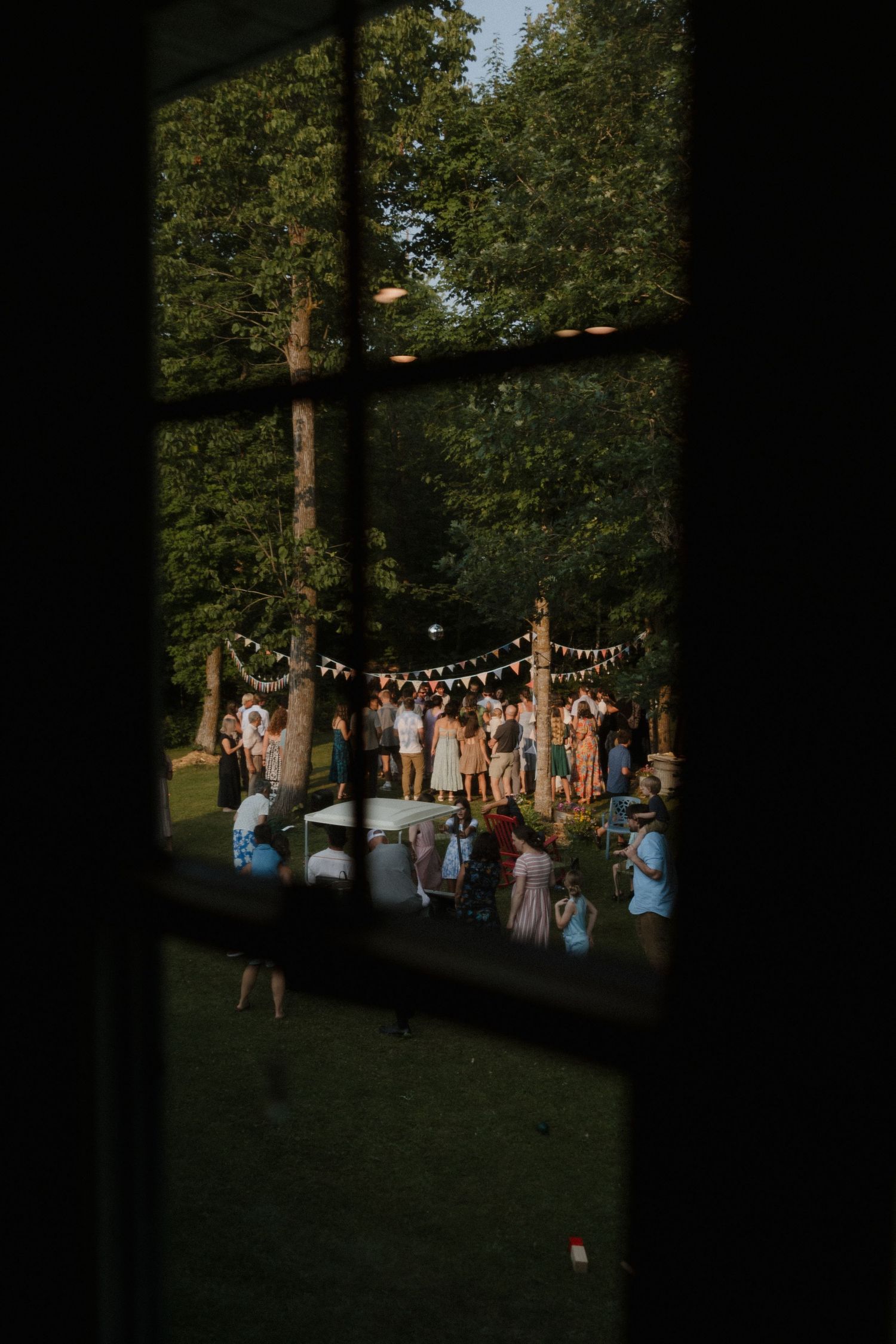 Evening wedding reception with guests mingling under string lights in a wooded setting.
