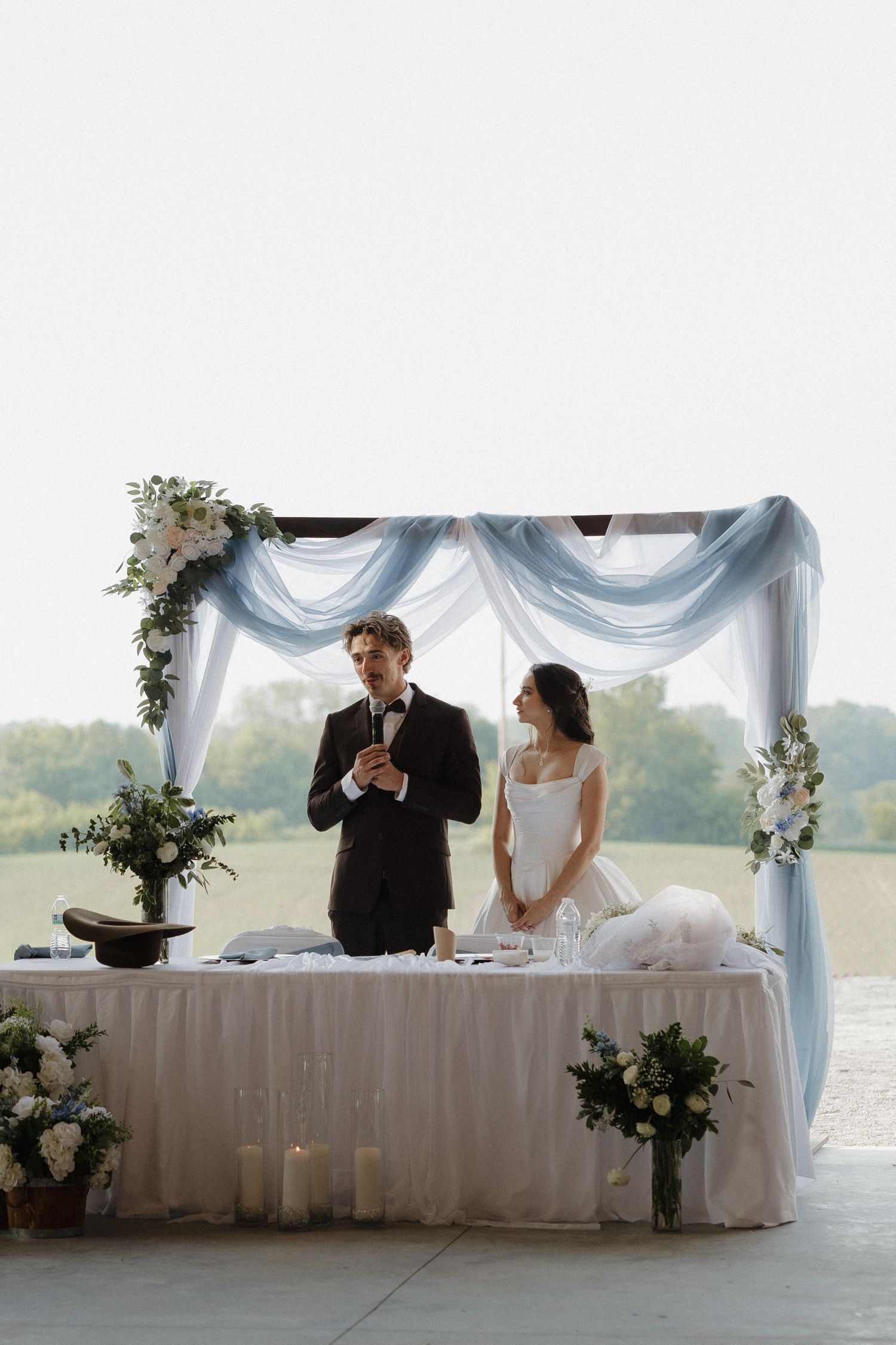 Couple seated at elegant white draped head table during outdoor wedding reception.