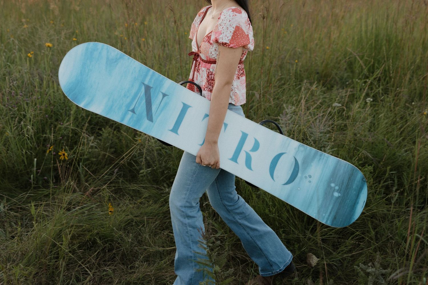 Young woman carrying a blue Nitro snowboard through a lush green grassy field outdoors.