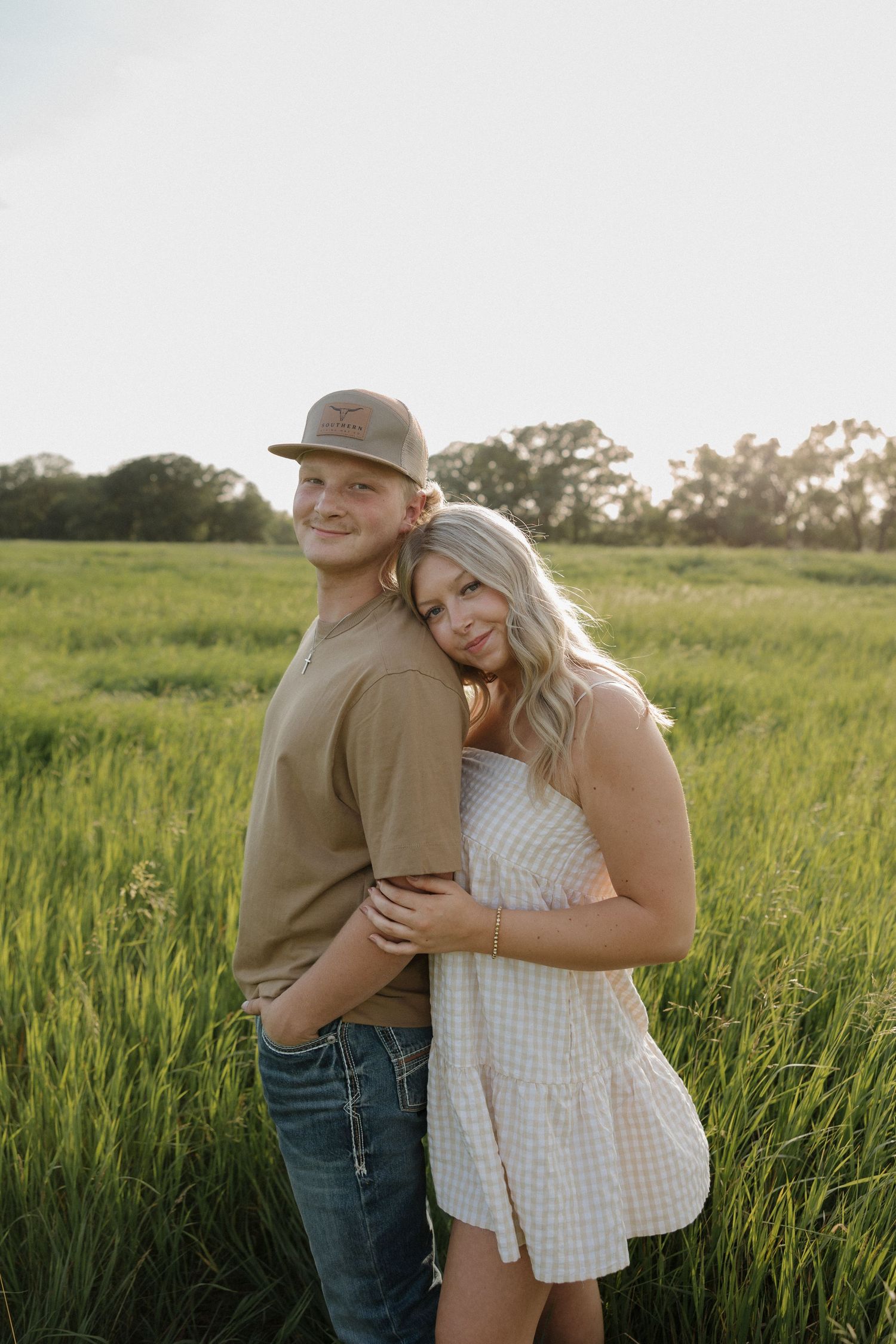 Couple posing in green field at sunset, woman leaning on man wearing cap and tan shirt.