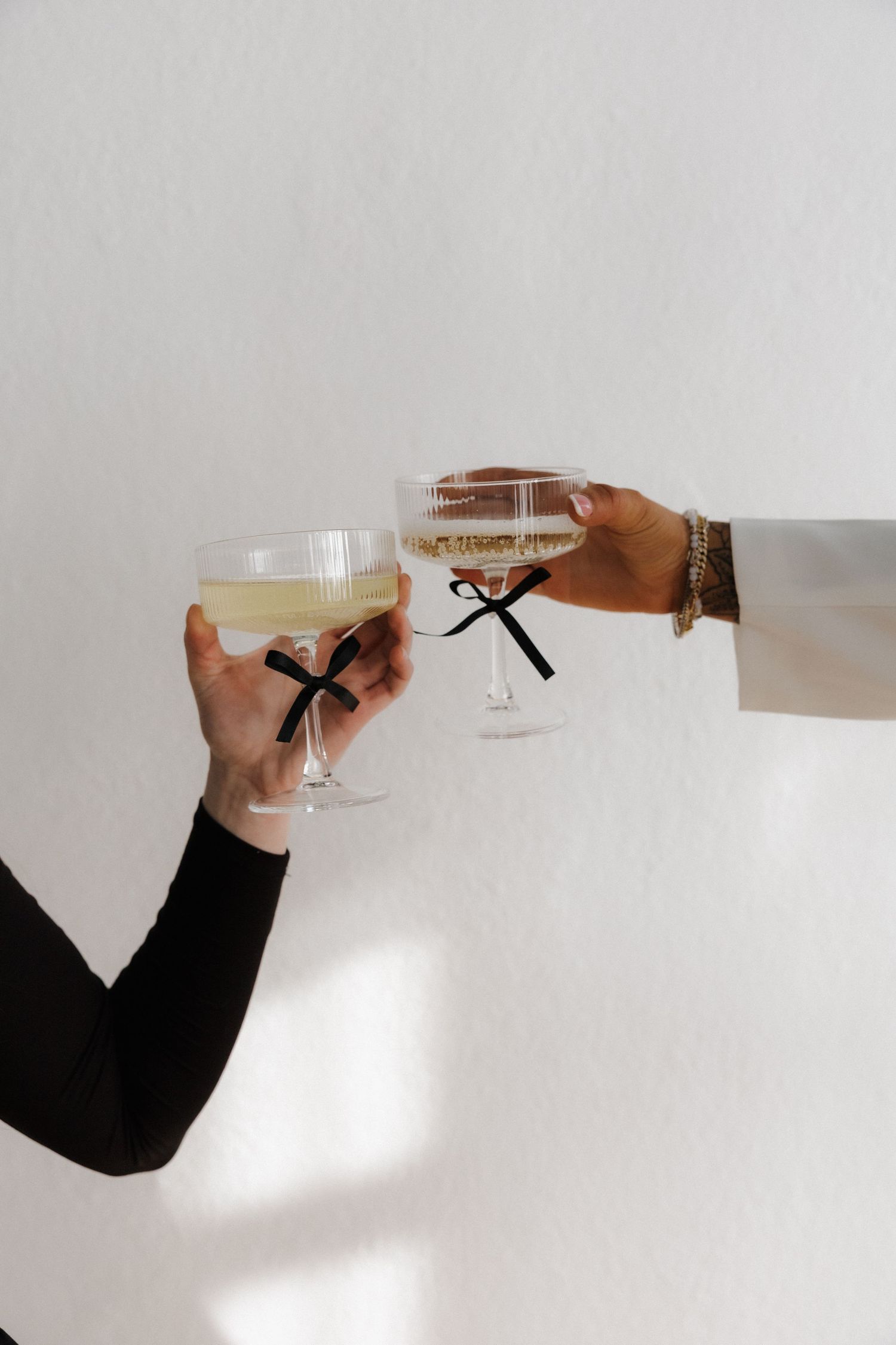 Two hands toasting with champagne coupe glasses adorned with black ribbon bows.