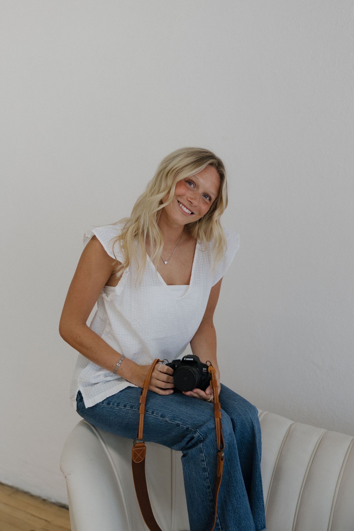 Blonde woman smiling while holding a DSLR camera seated on a white chair in a studio.
