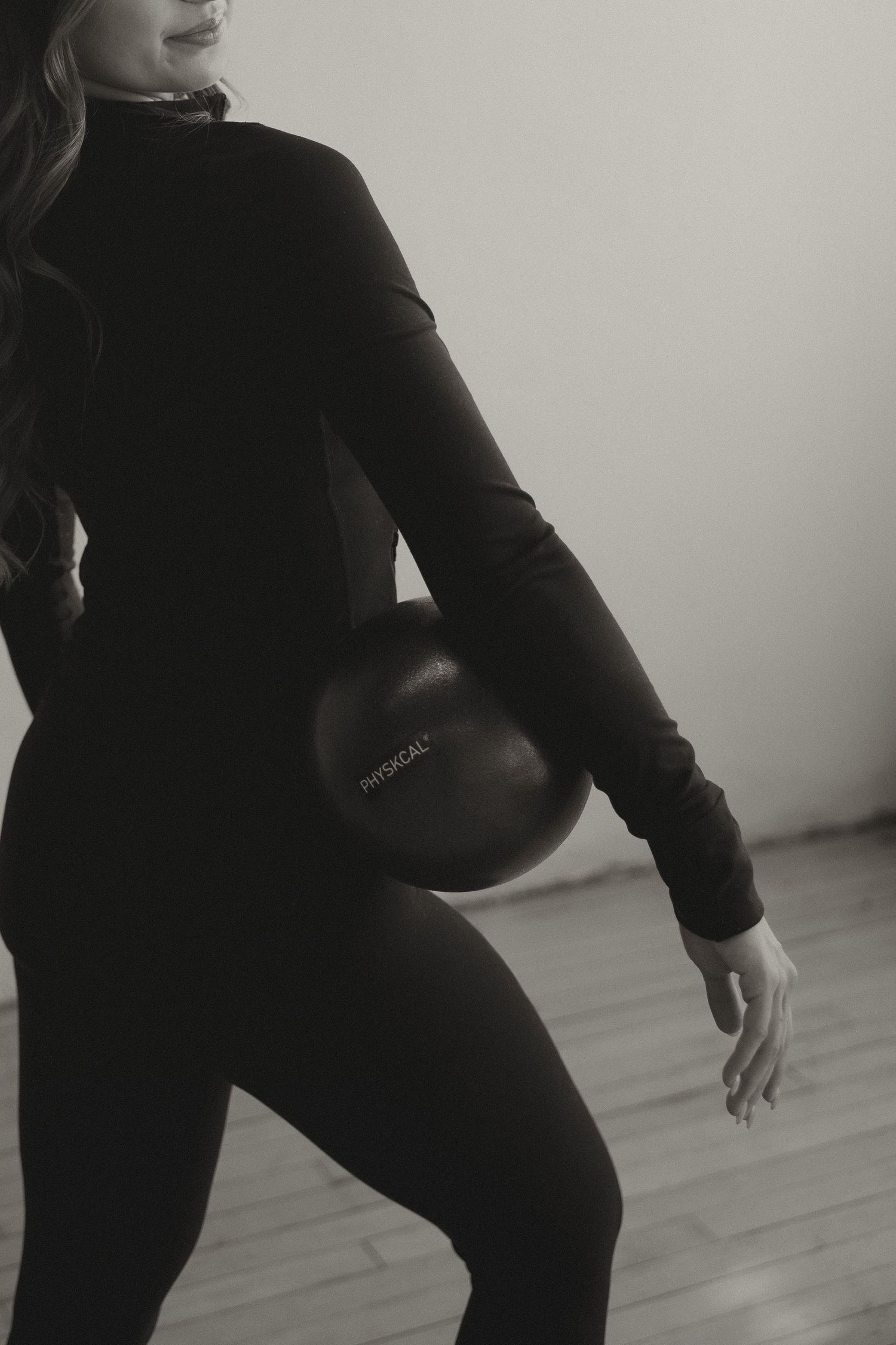 Black and white photo of woman in black athletic outfit holding a Physkcal exercise ball at her side.