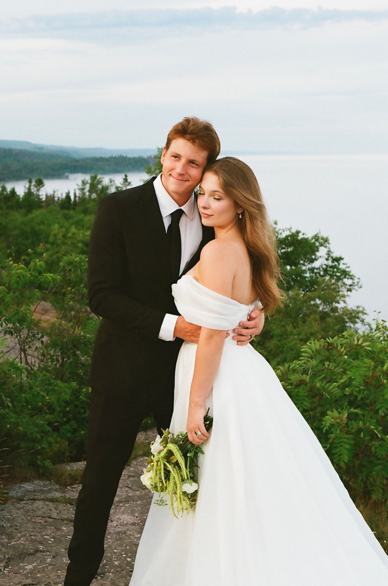 Bride and groom embrace on a scenic overlook, she in an off-shoulder gown holding a bouquet, he in a black suit.