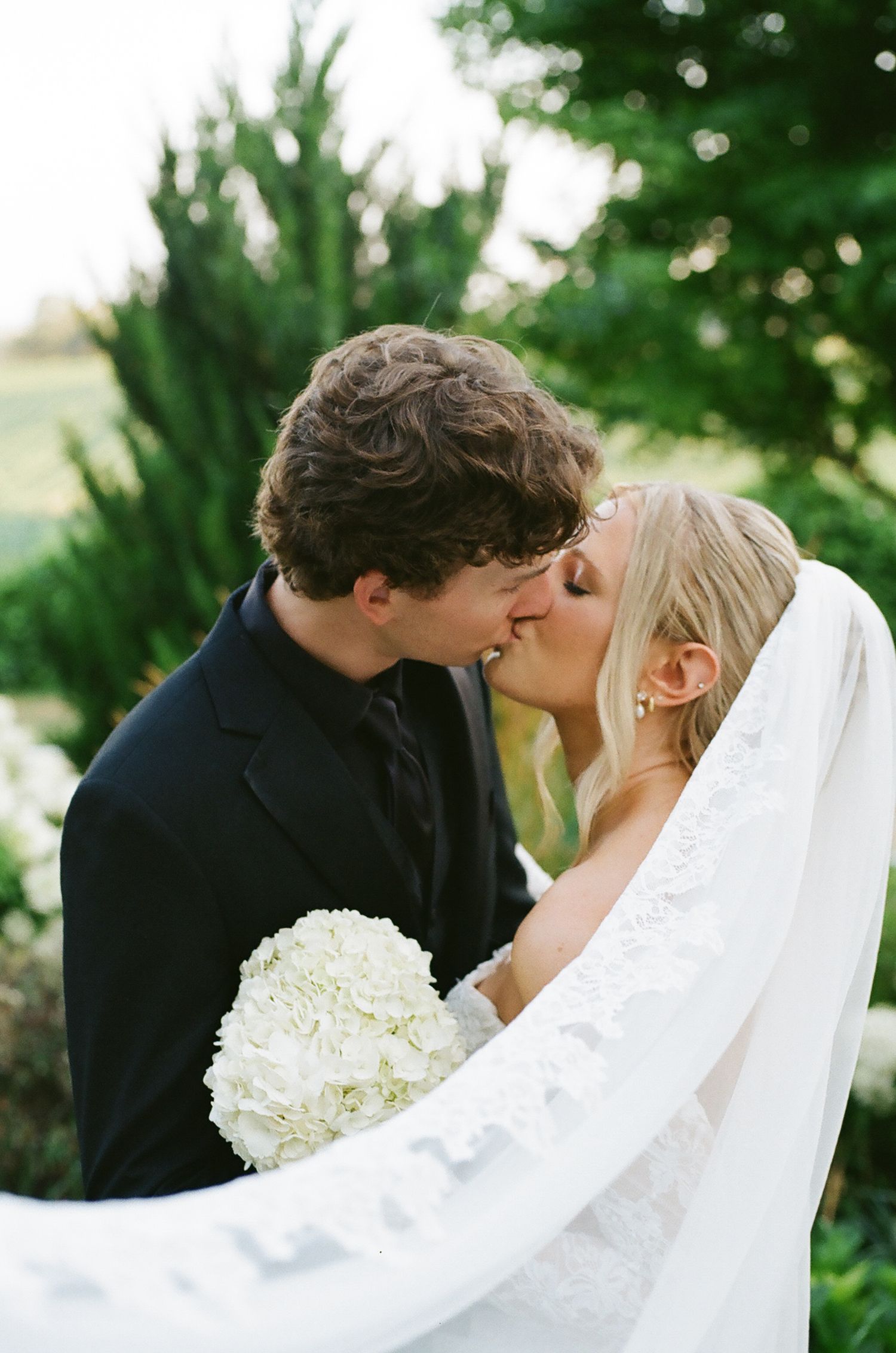 Bride and groom share a romantic kiss, bride holding white hydrangea bouquet with veil flowing, surrounded by lush greenery.