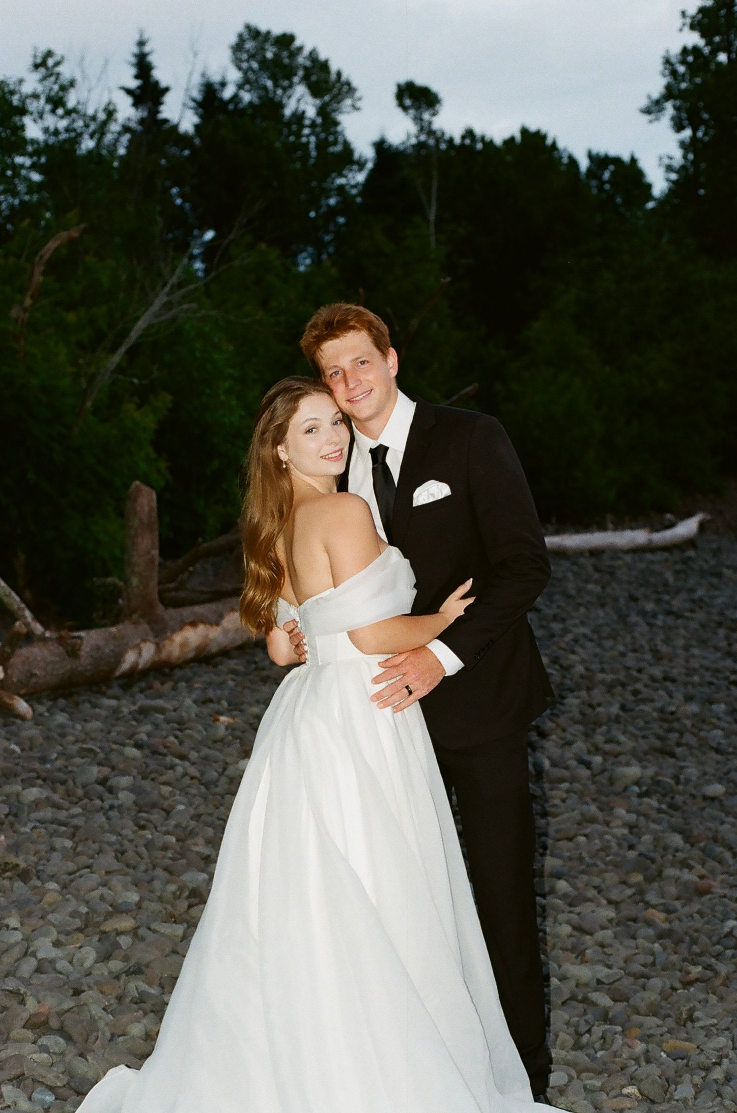 A bride in a white off-shoulder gown and groom in a black tuxedo embrace on a rocky beach surrounded by trees.