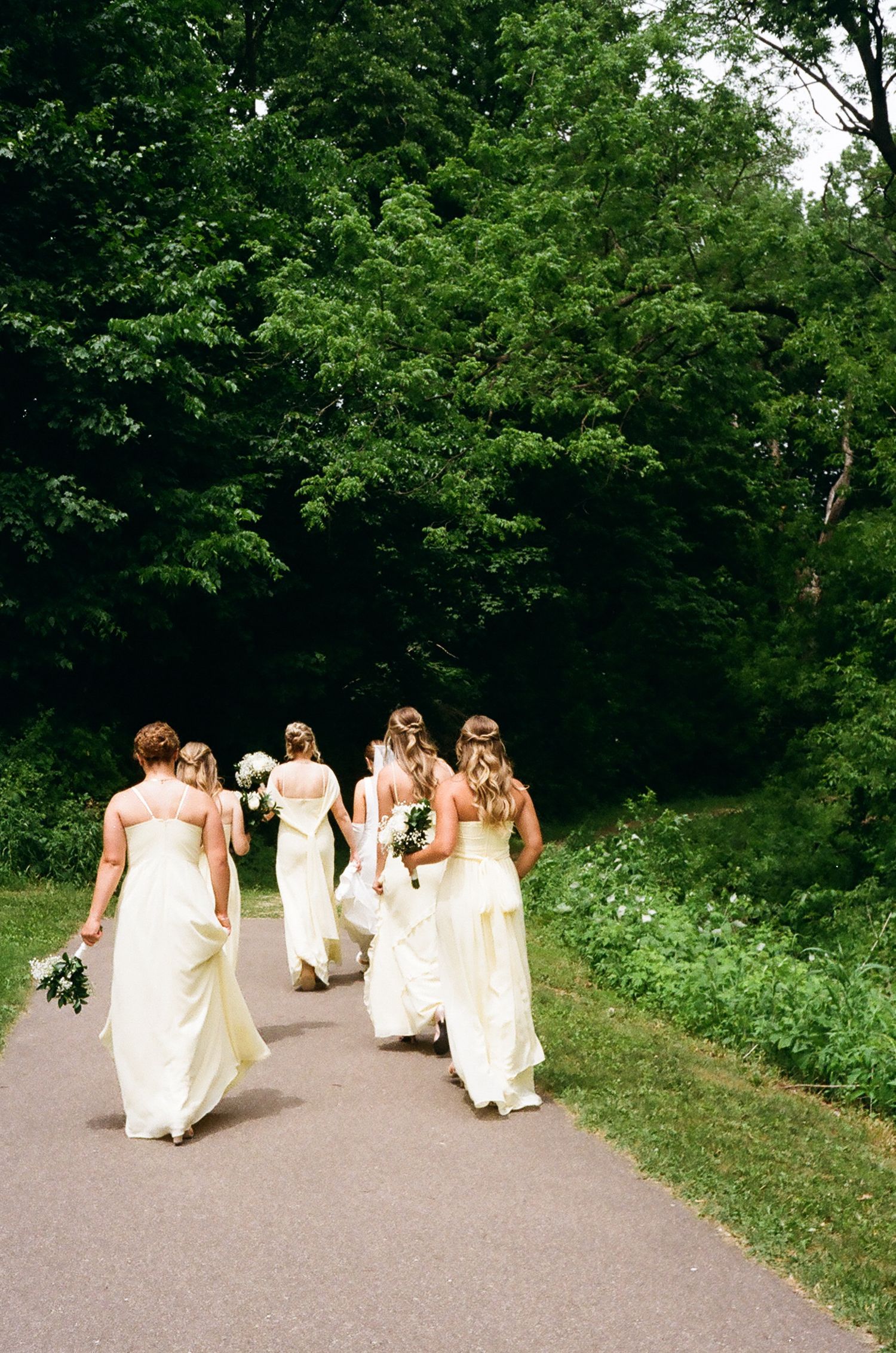 Bride and bridesmaids in white gowns walking down a tree-lined path outdoors.