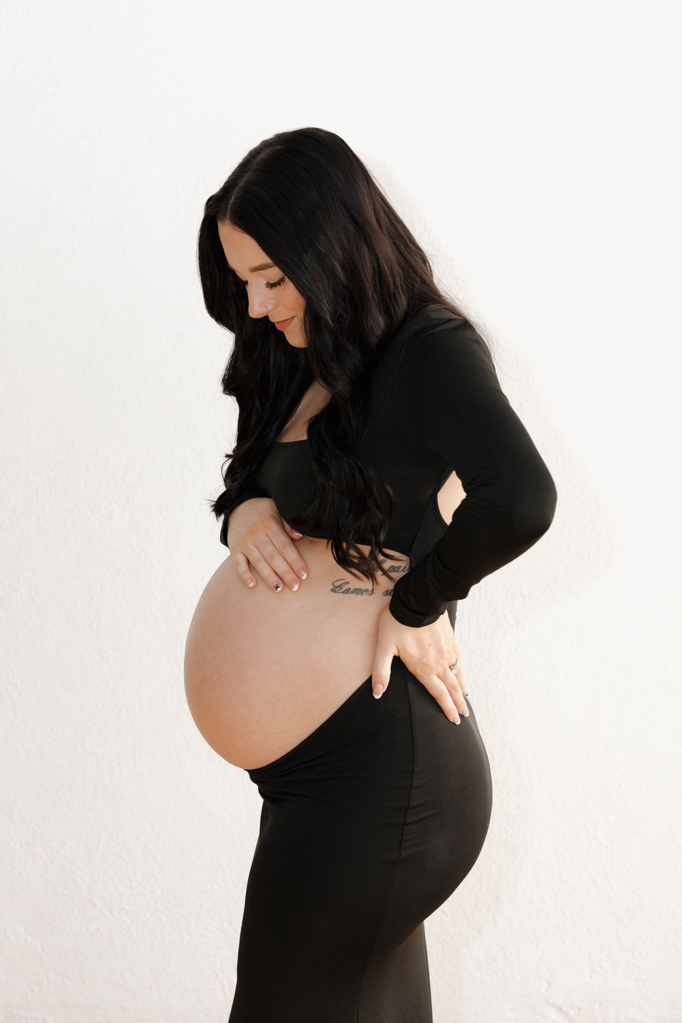 Pregnant woman in black outfit cradling large baby bump while looking down against white wall.