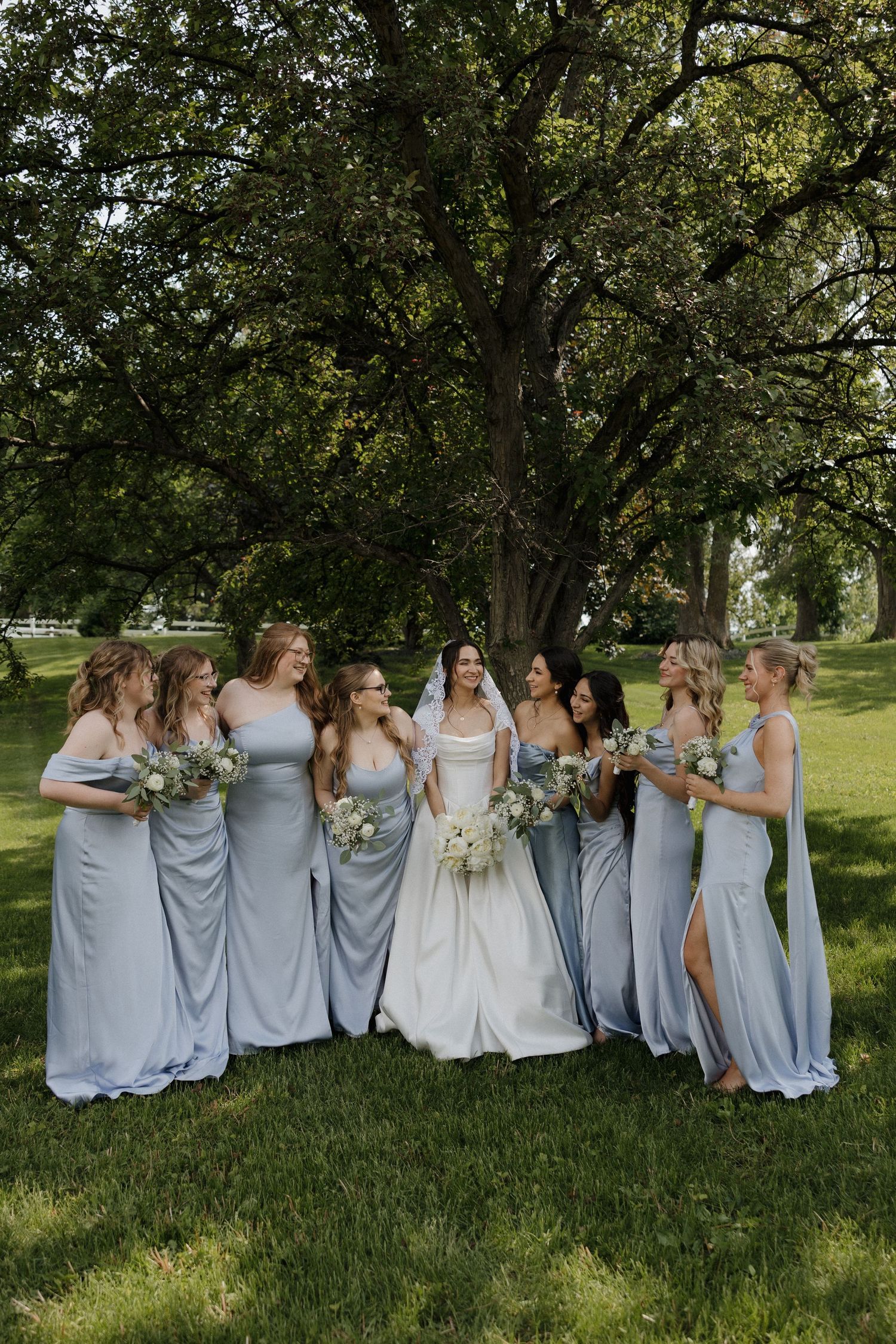 Wedding party wearing light blue bridesmaid dresses posing together outdoors under trees.
