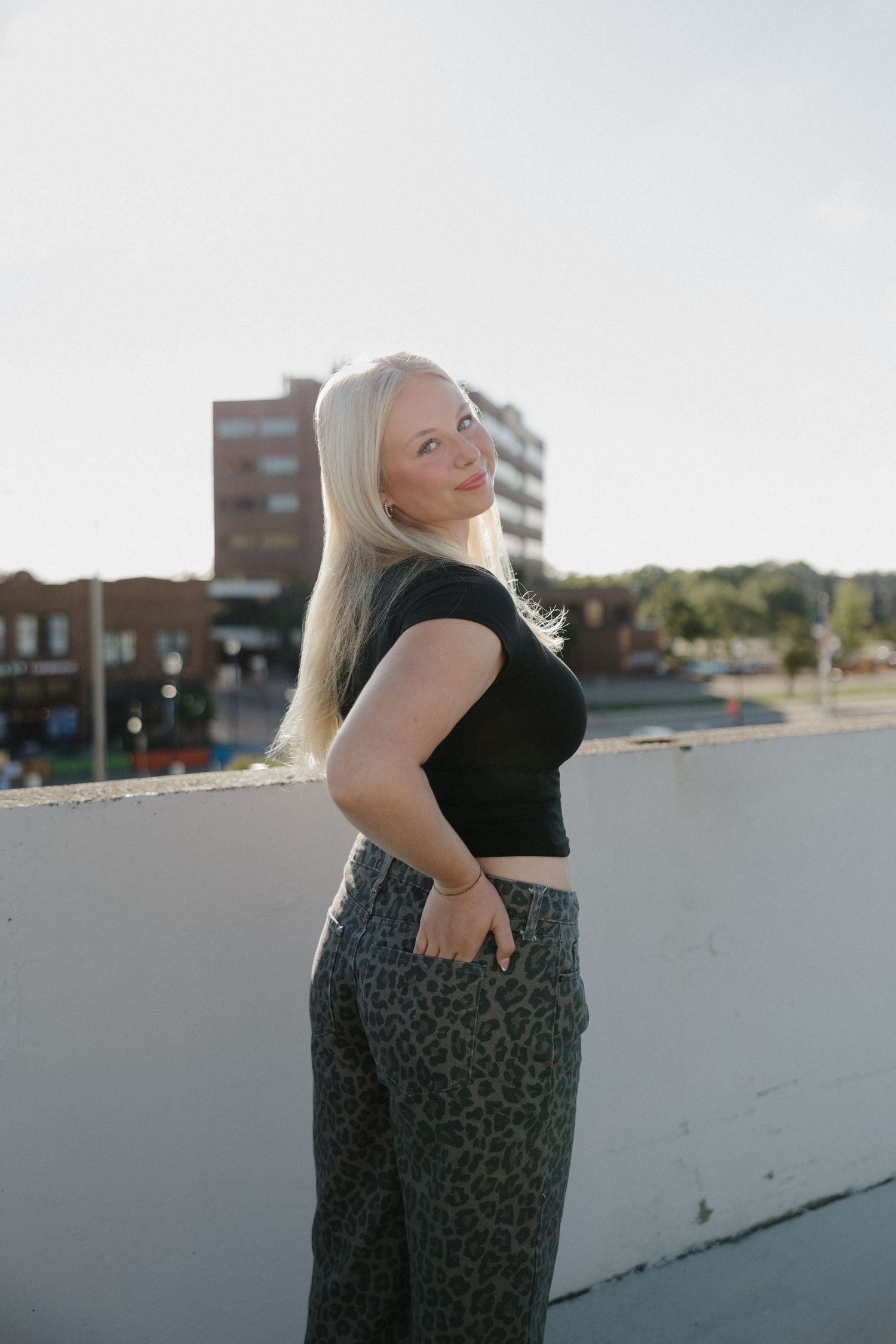 Blonde woman in black crop top and leopard print pants posing on a rooftop with urban buildings behind her.