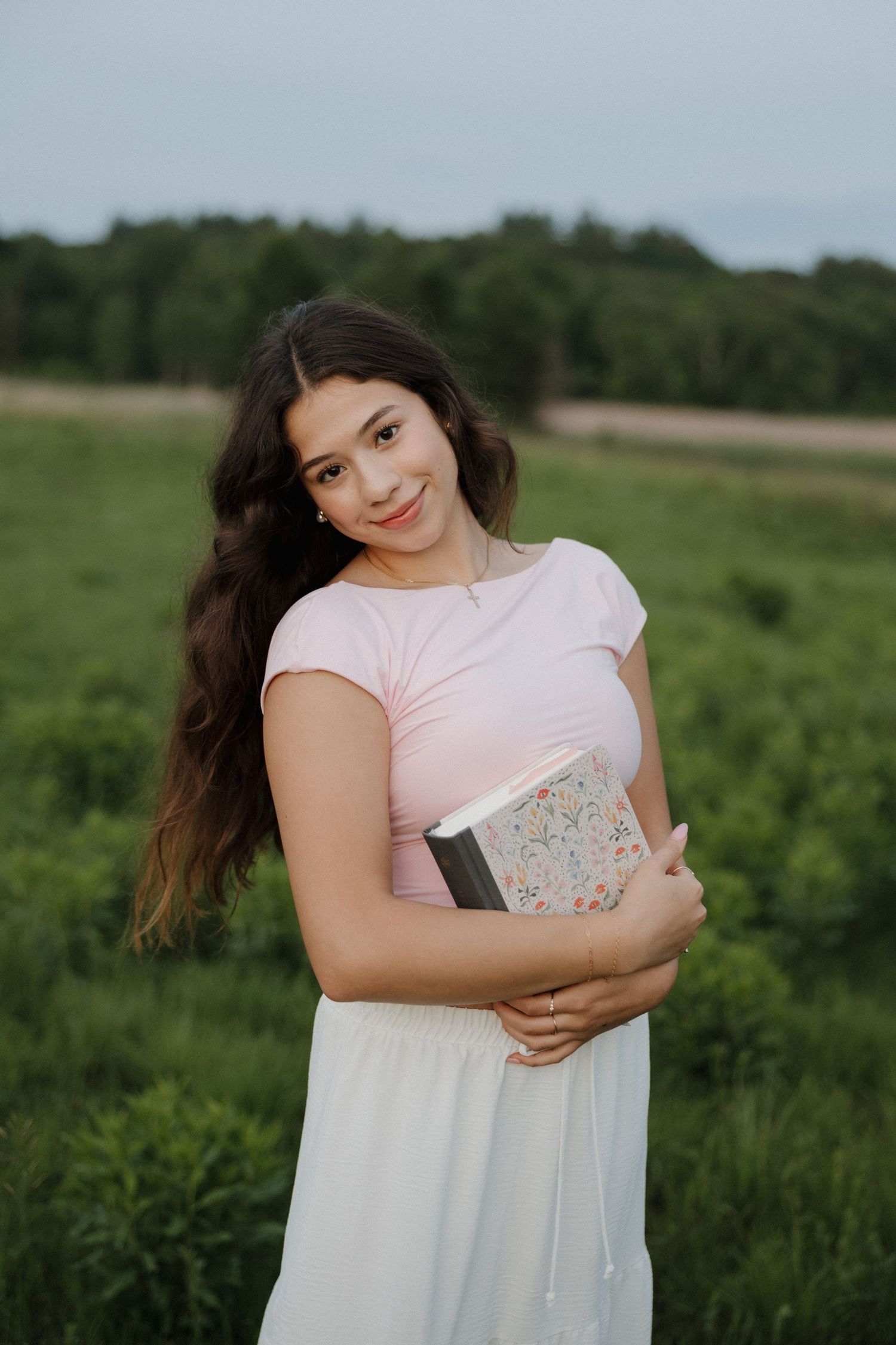Young woman in pink top and white skirt hugging a book while smiling in a green field.
