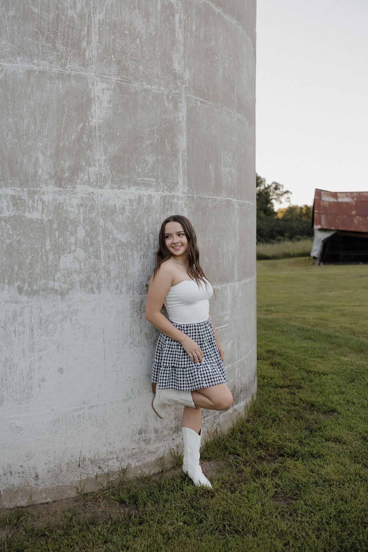 Young woman in white tube top, plaid skirt, and white cowboy boots leaning against a concrete silo wall.
