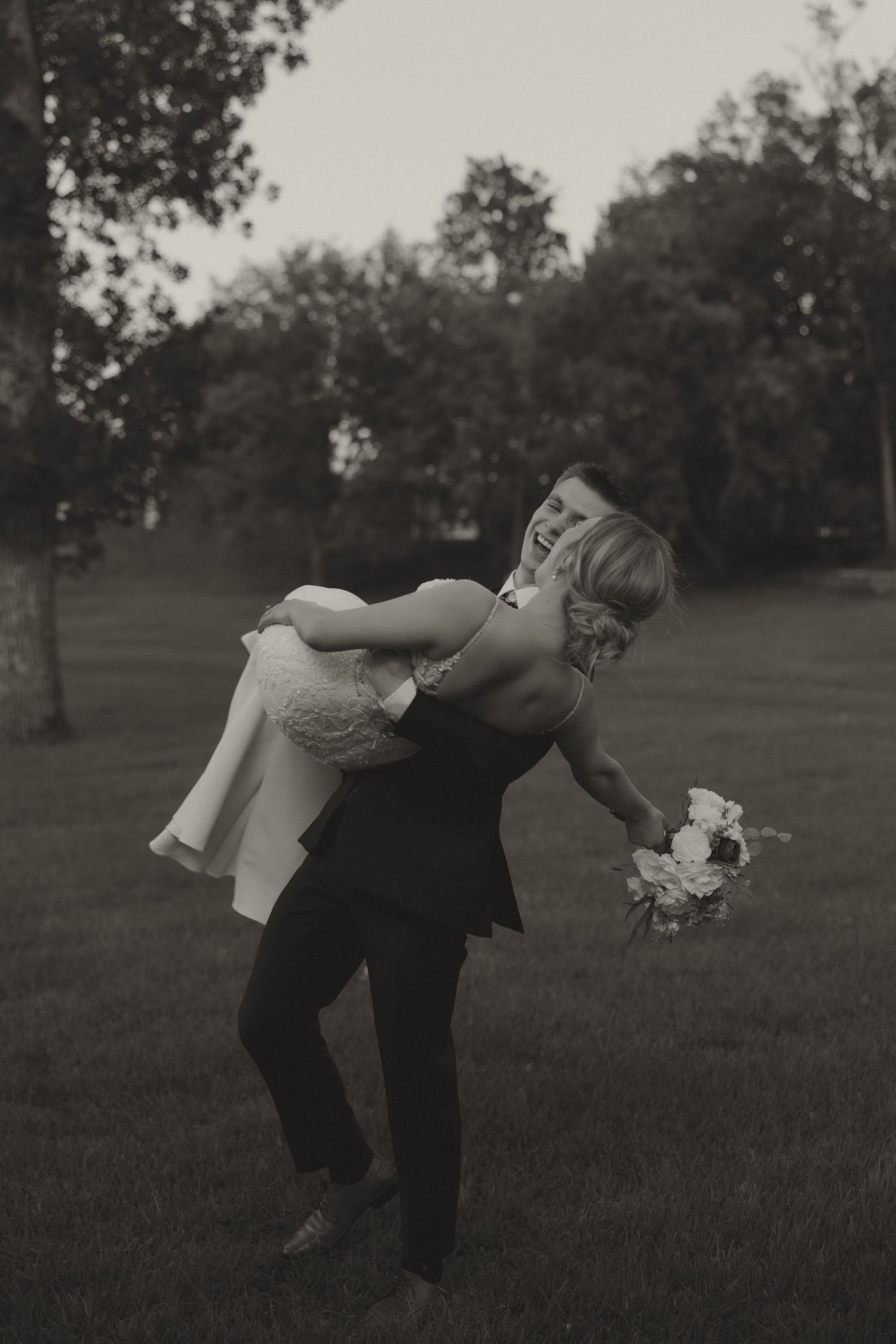 Groom playfully carries laughing bride in a black and white outdoor wedding portrait.
