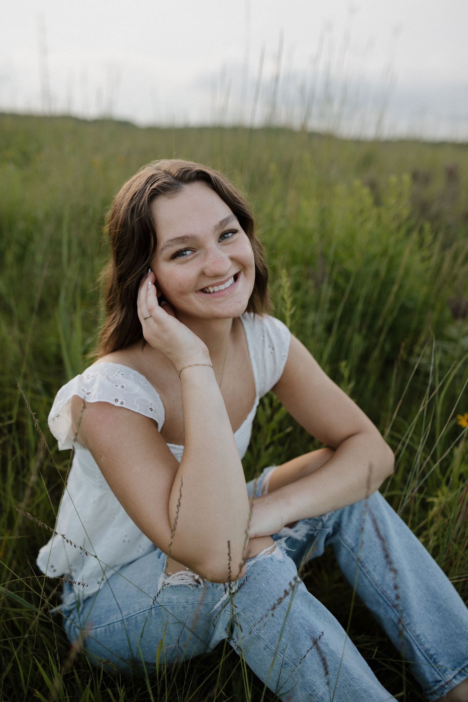 Smiling young woman in a white top and ripped jeans sitting in tall green grass outdoors.