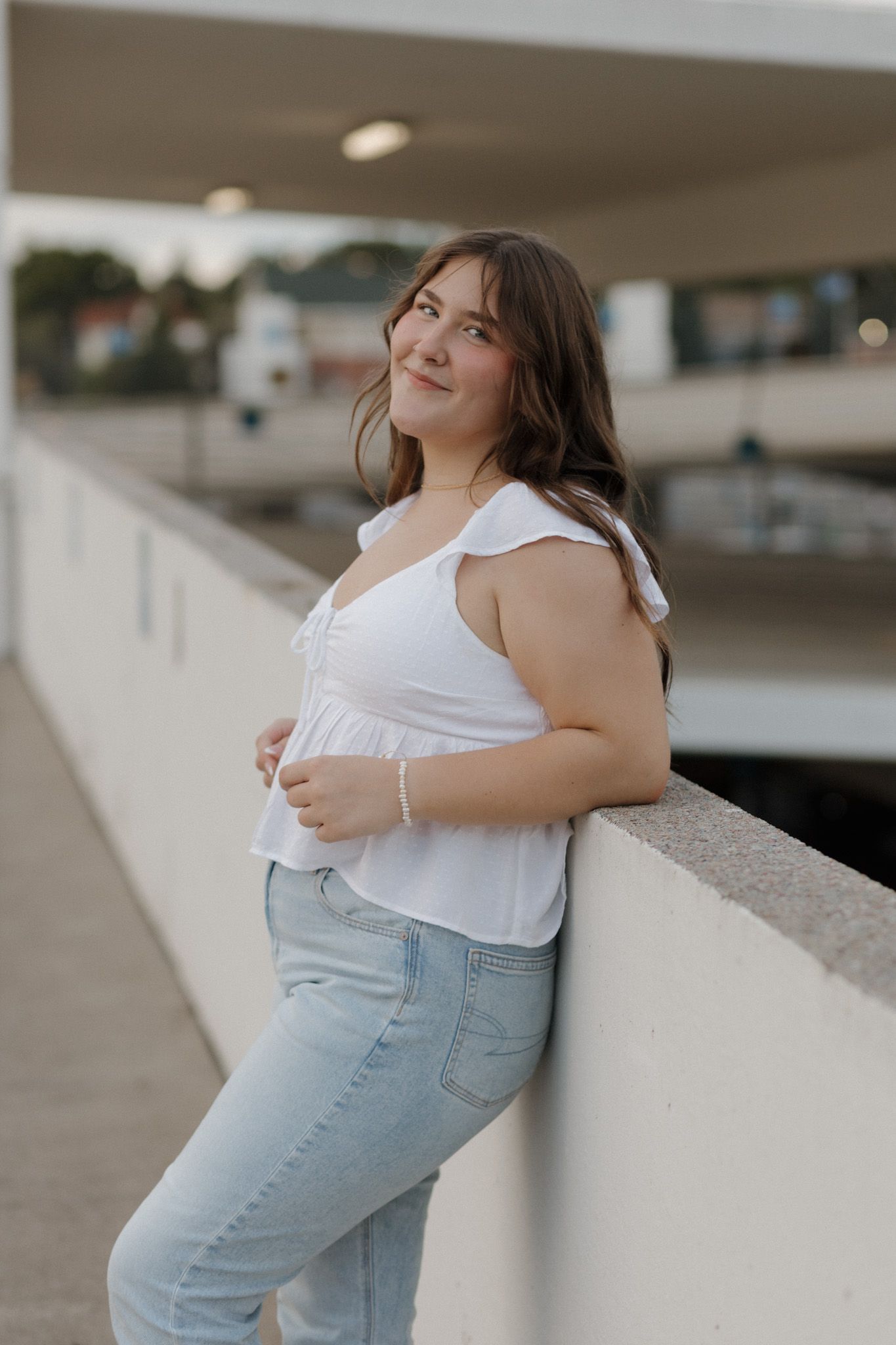 Young woman in white ruffled top and jeans leaning against concrete wall at sunset.