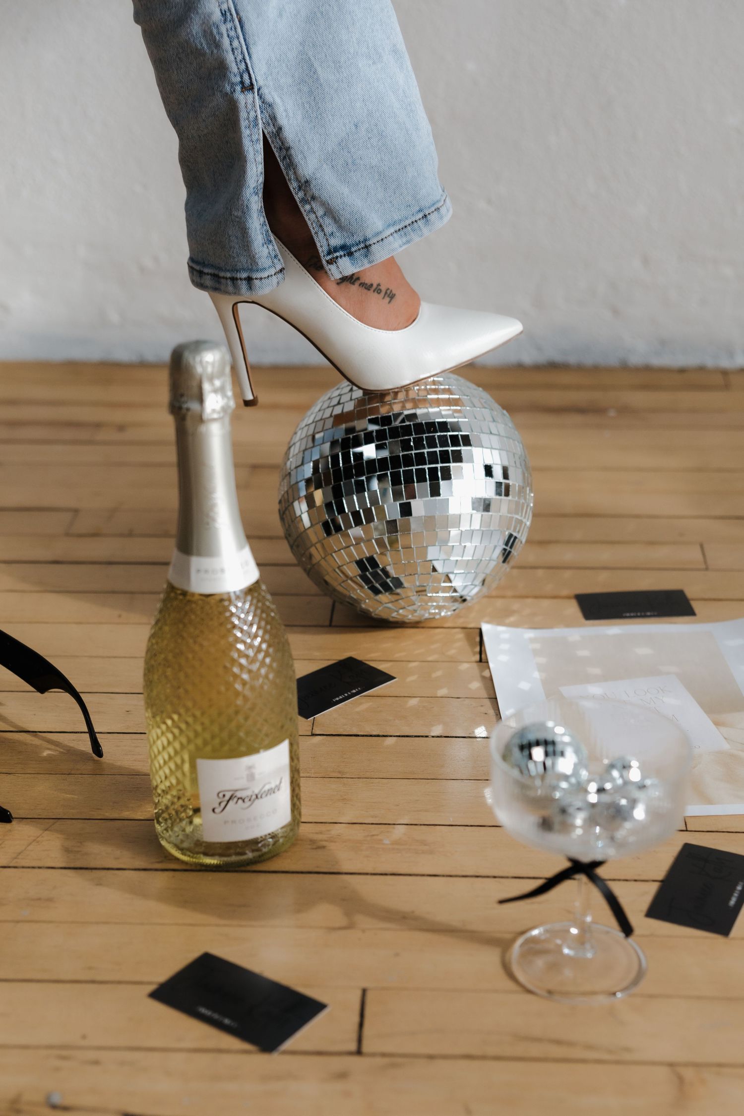 White stiletto heel resting on a disco ball beside a champagne bottle and cocktail glass on a wooden floor.