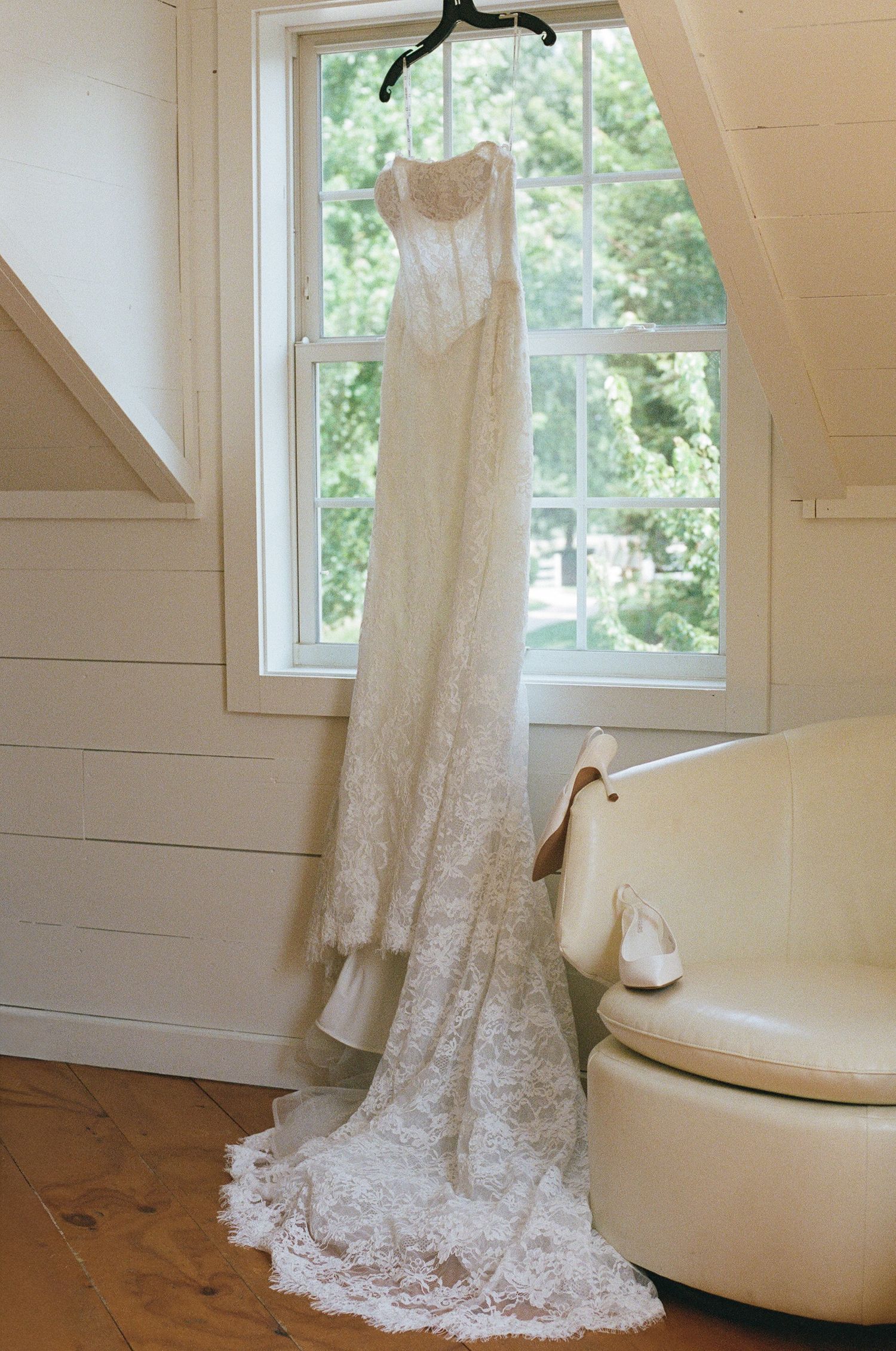 Elegant lace wedding dress hanging in a sunlit window with a cathedral train pooling on hardwood floors beside a cream chair.