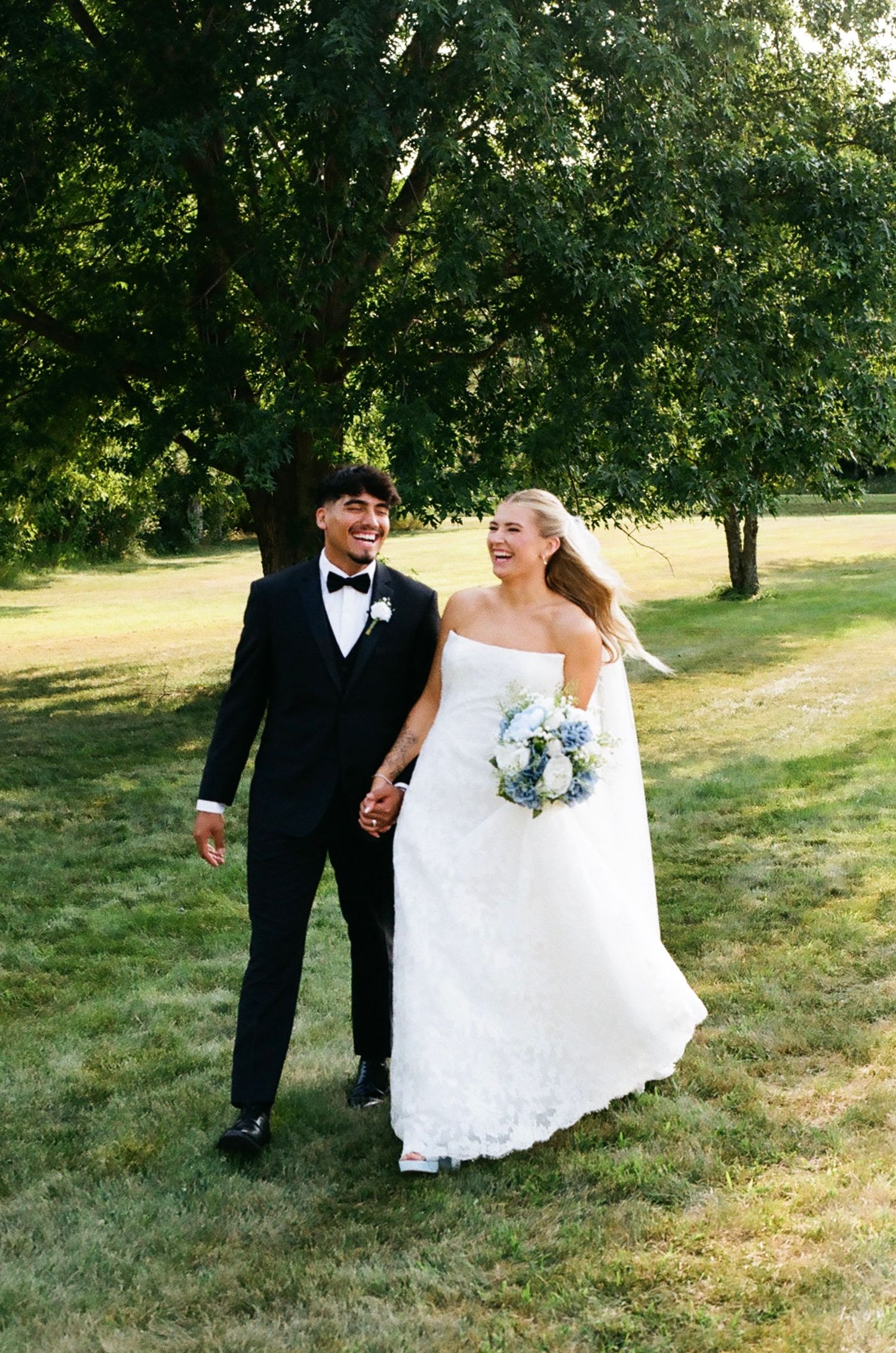 A smiling bride in a white strapless gown and groom in a black tuxedo walk hand-in-hand on a green lawn.