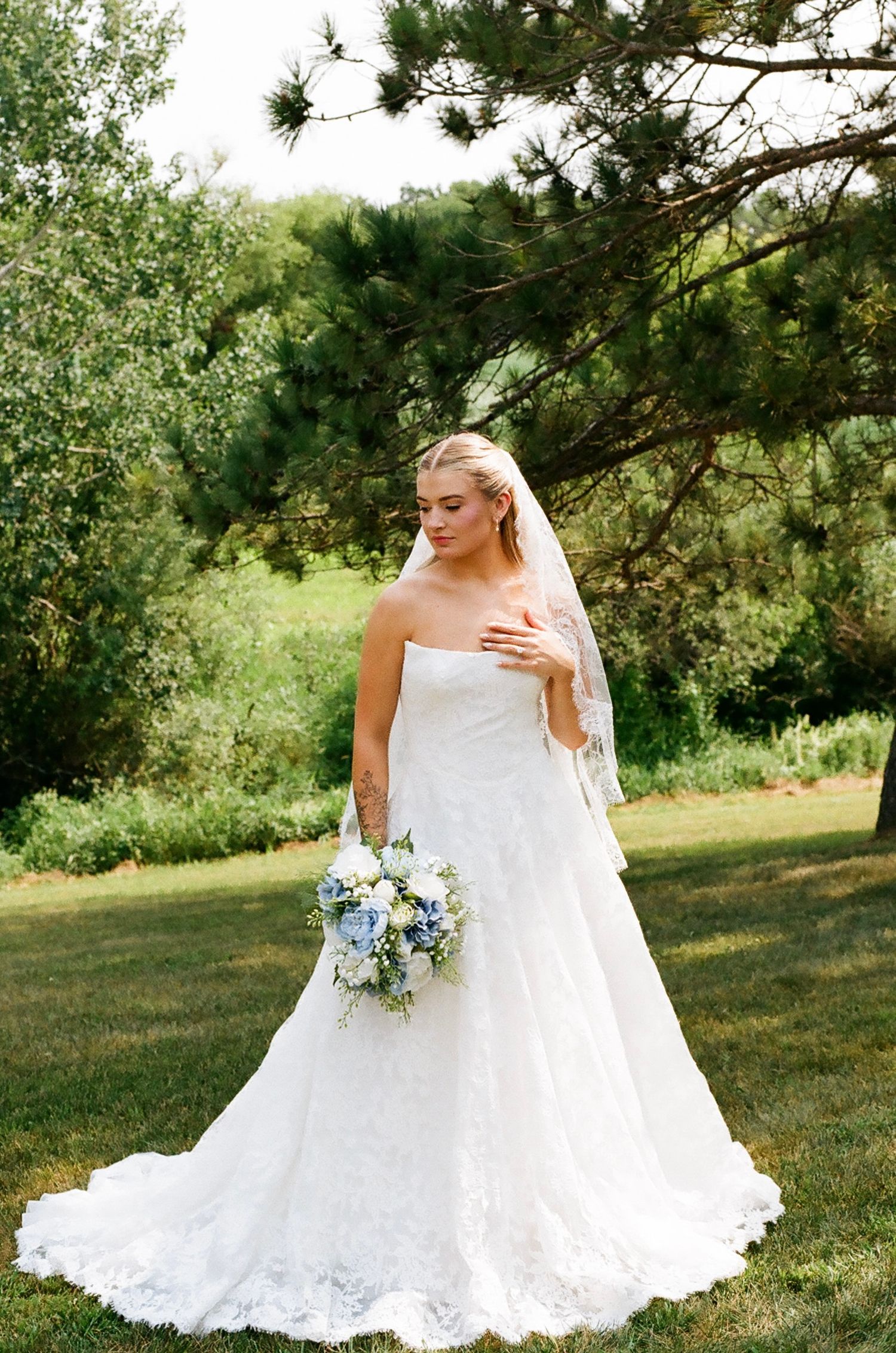Bride in strapless lace wedding gown with veil holding blue and white bouquet outdoors.