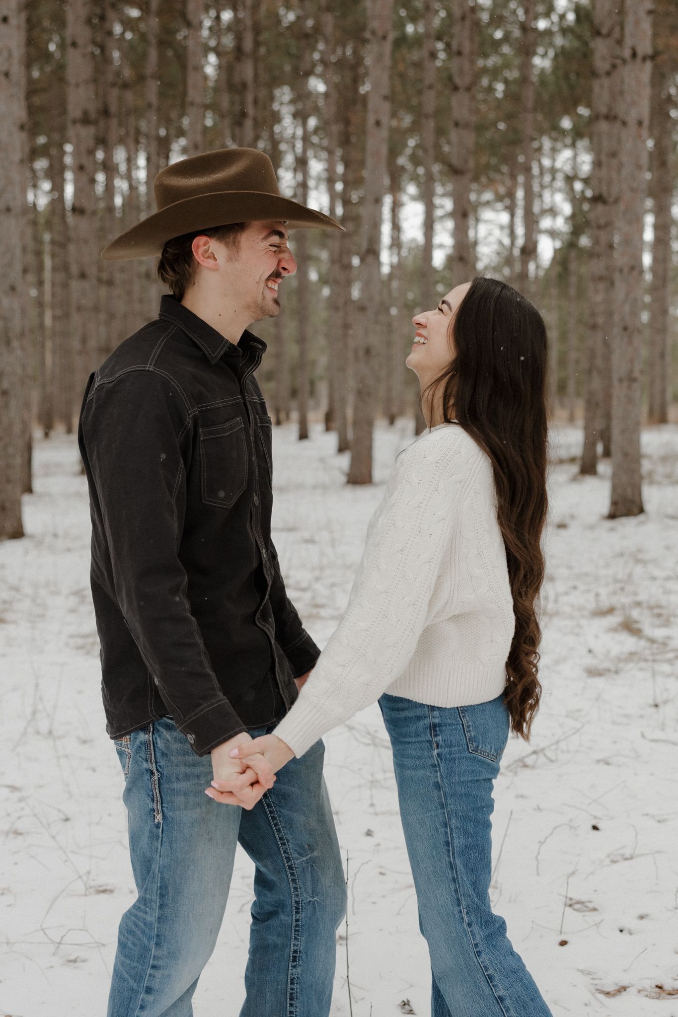 Laughing couple holding hands in snowy pine forest, man wearing brown cowboy hat and black jacket.
