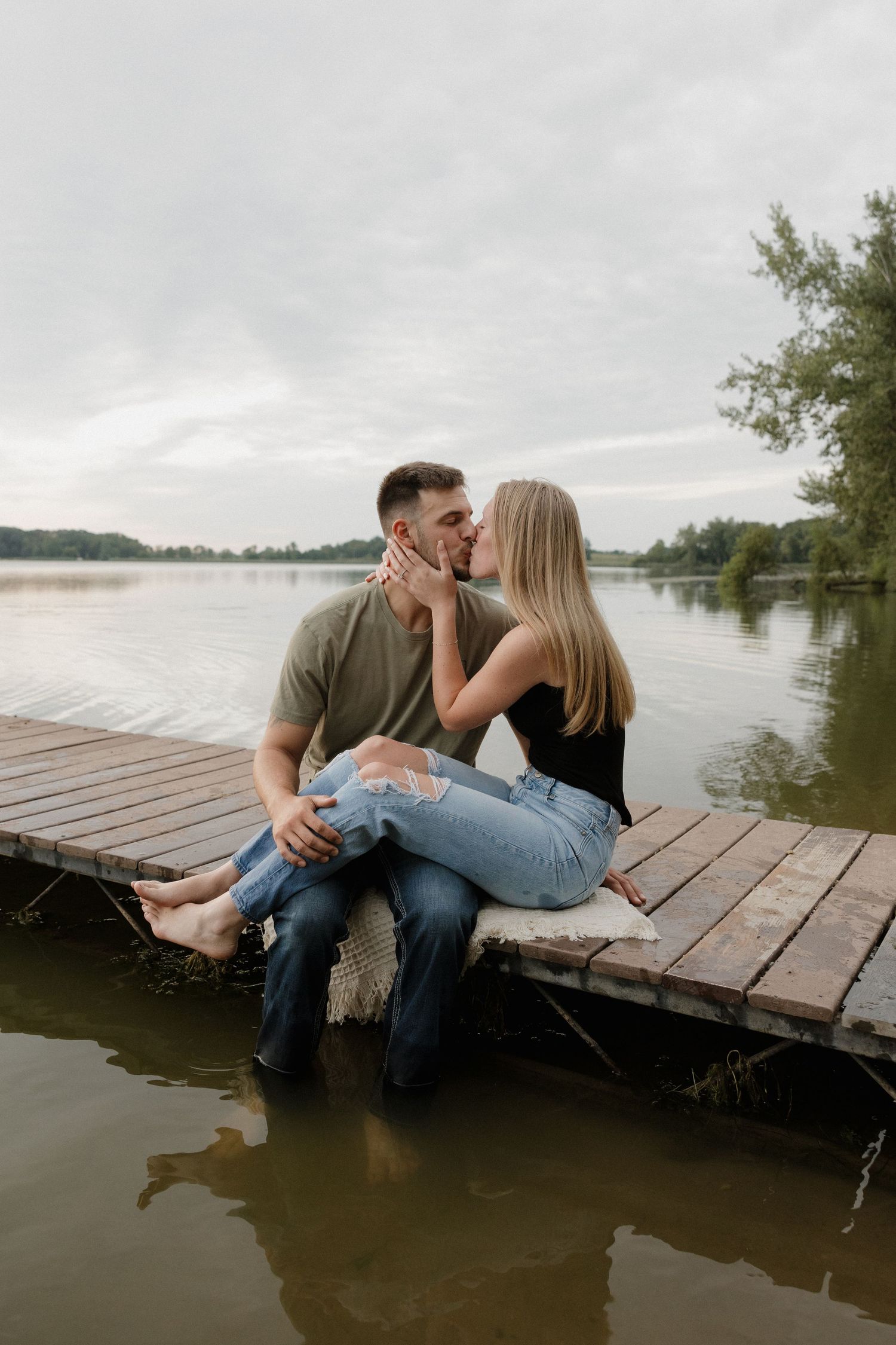 Couple sharing a kiss while sitting on a wooden dock over a calm lake.