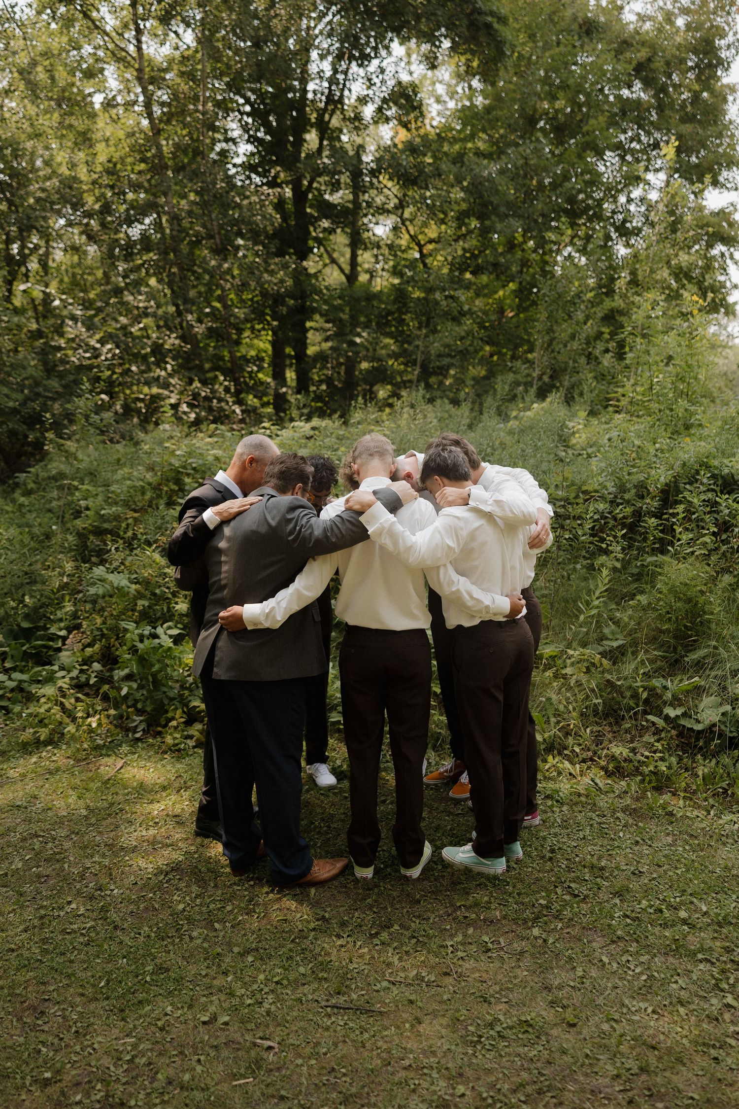 Wedding party in formal attire forms a huddle in a green outdoor setting.