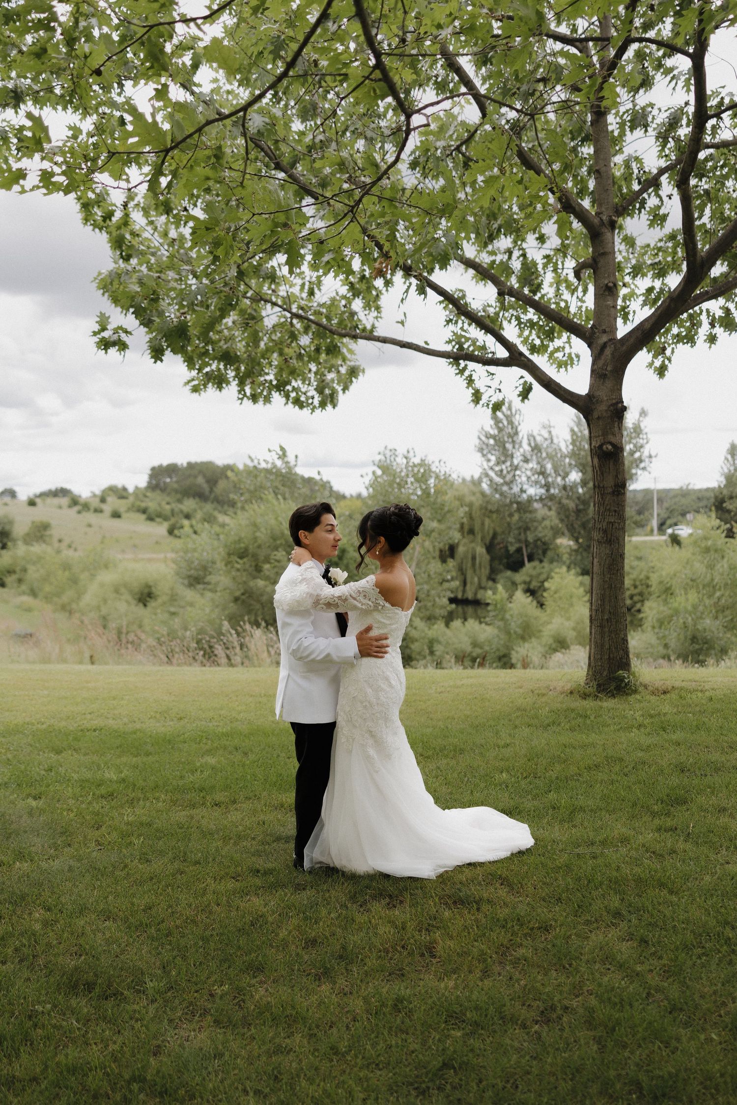 A romantic wedding couple shares a tender moment under a large tree in a scenic outdoor setting.