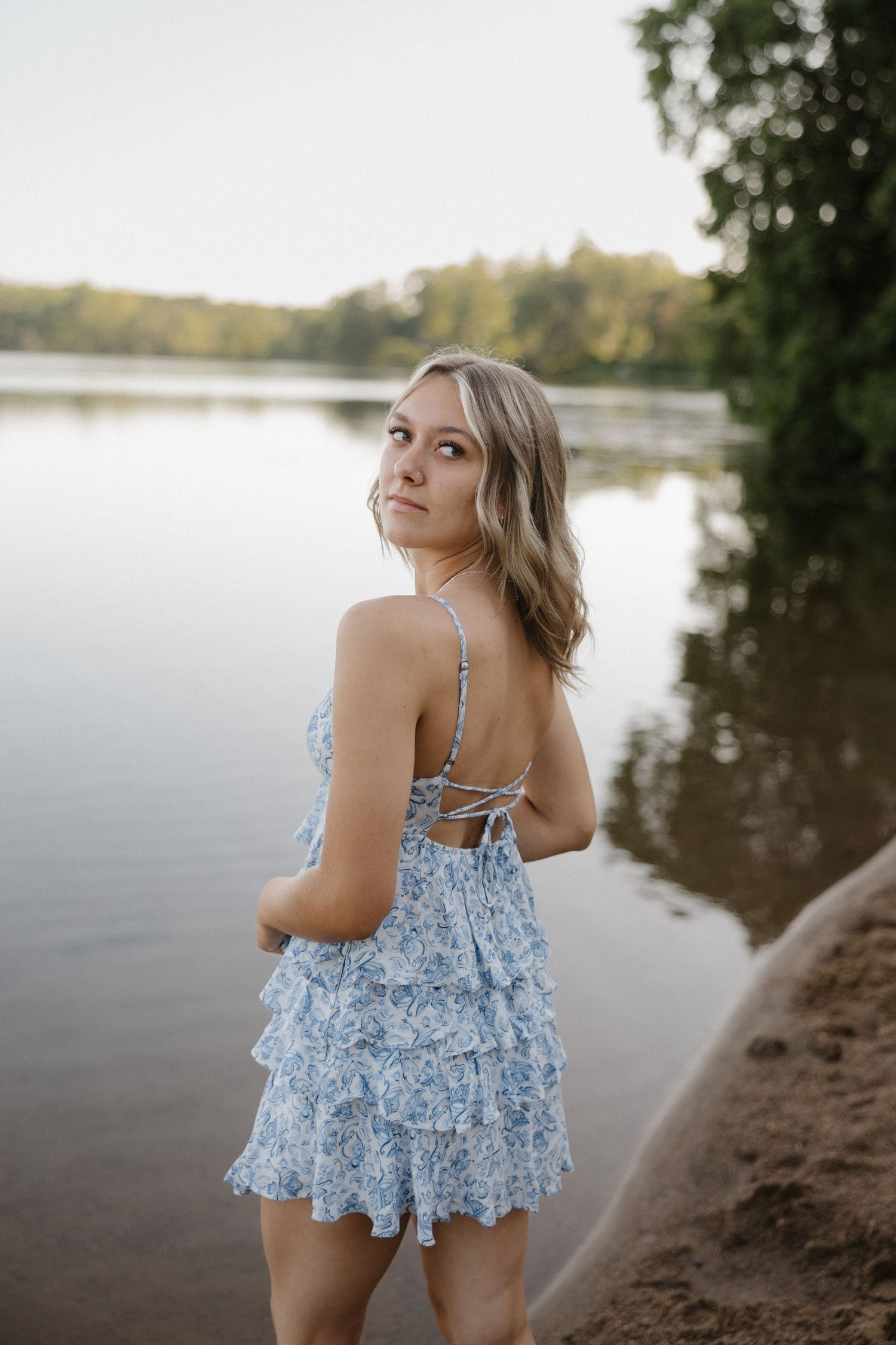 Young blonde woman in blue ruffled romper standing by calm lake at sunset.
