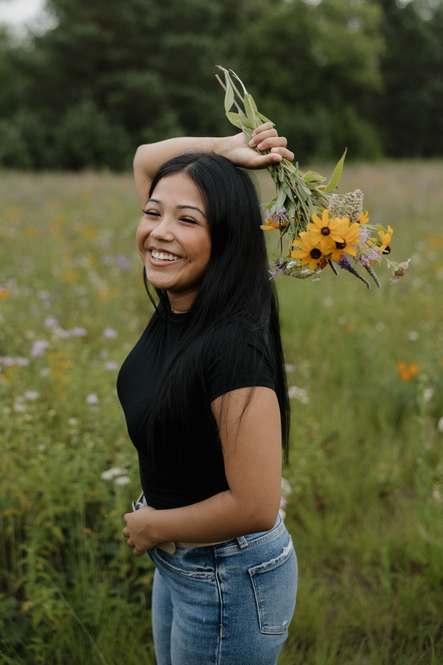 Smiling young woman holding wildflowers above her head in a colorful meadow, wearing a black tee and jeans.