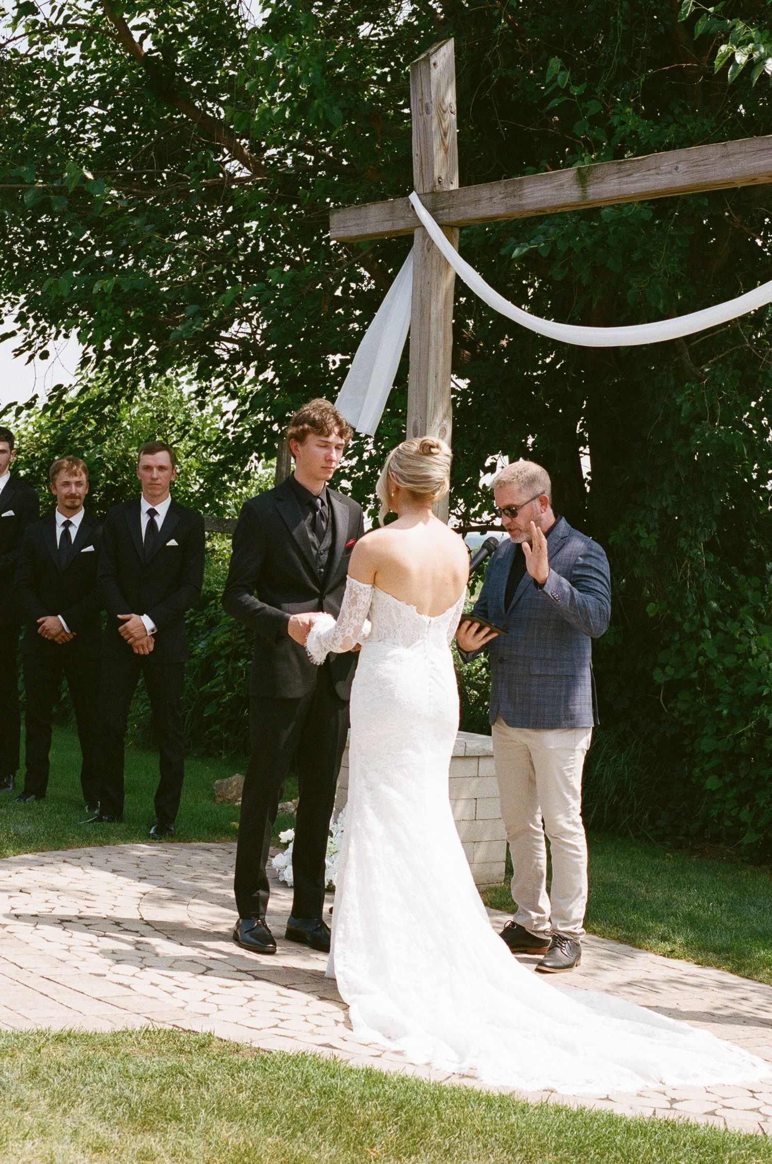 Outdoor wedding ceremony with bride and groom exchanging vows before a wooden cross, groomsmen standing nearby.