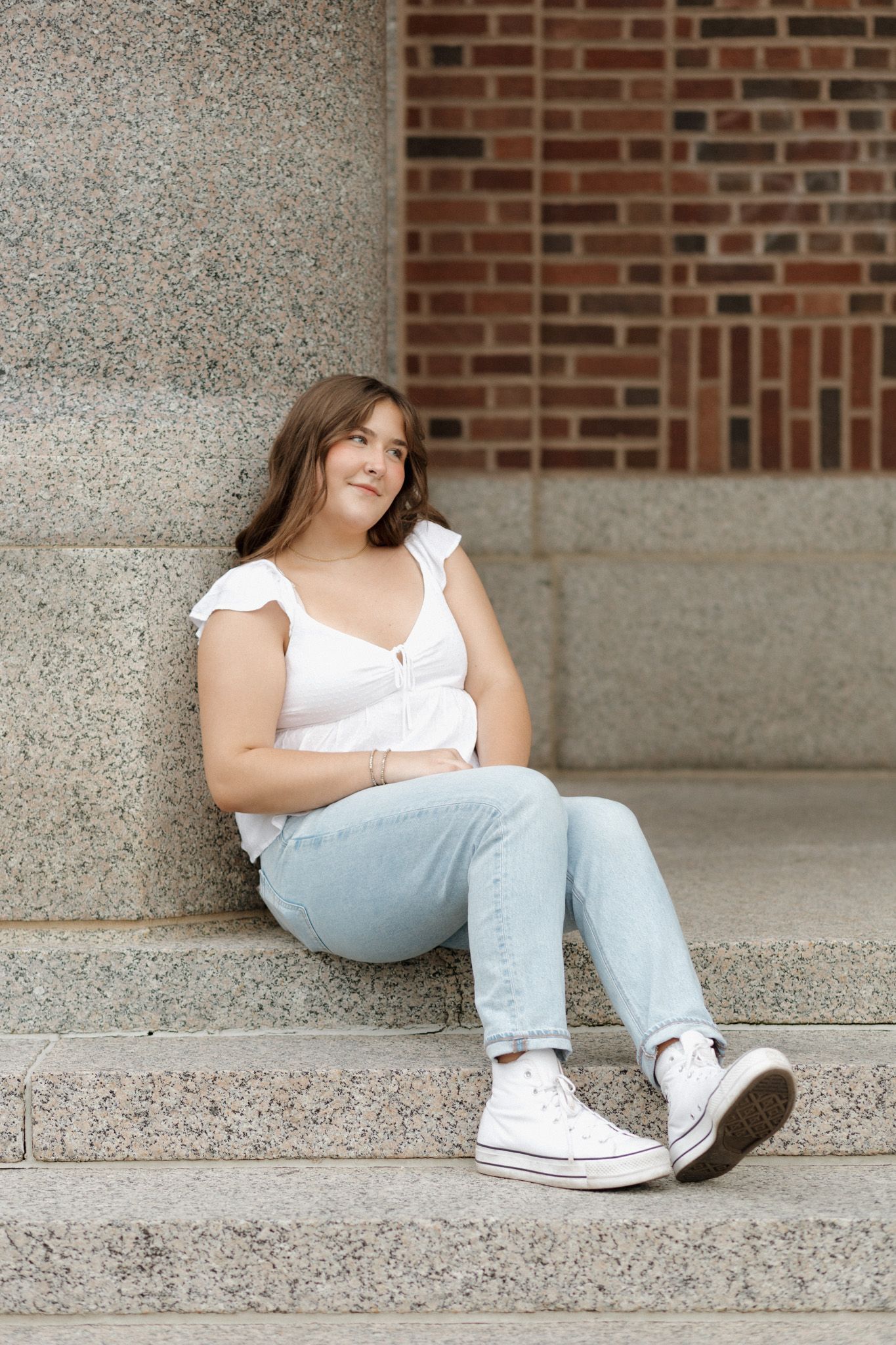 Teen girl in white top and light jeans sitting on stone steps near brick wall.