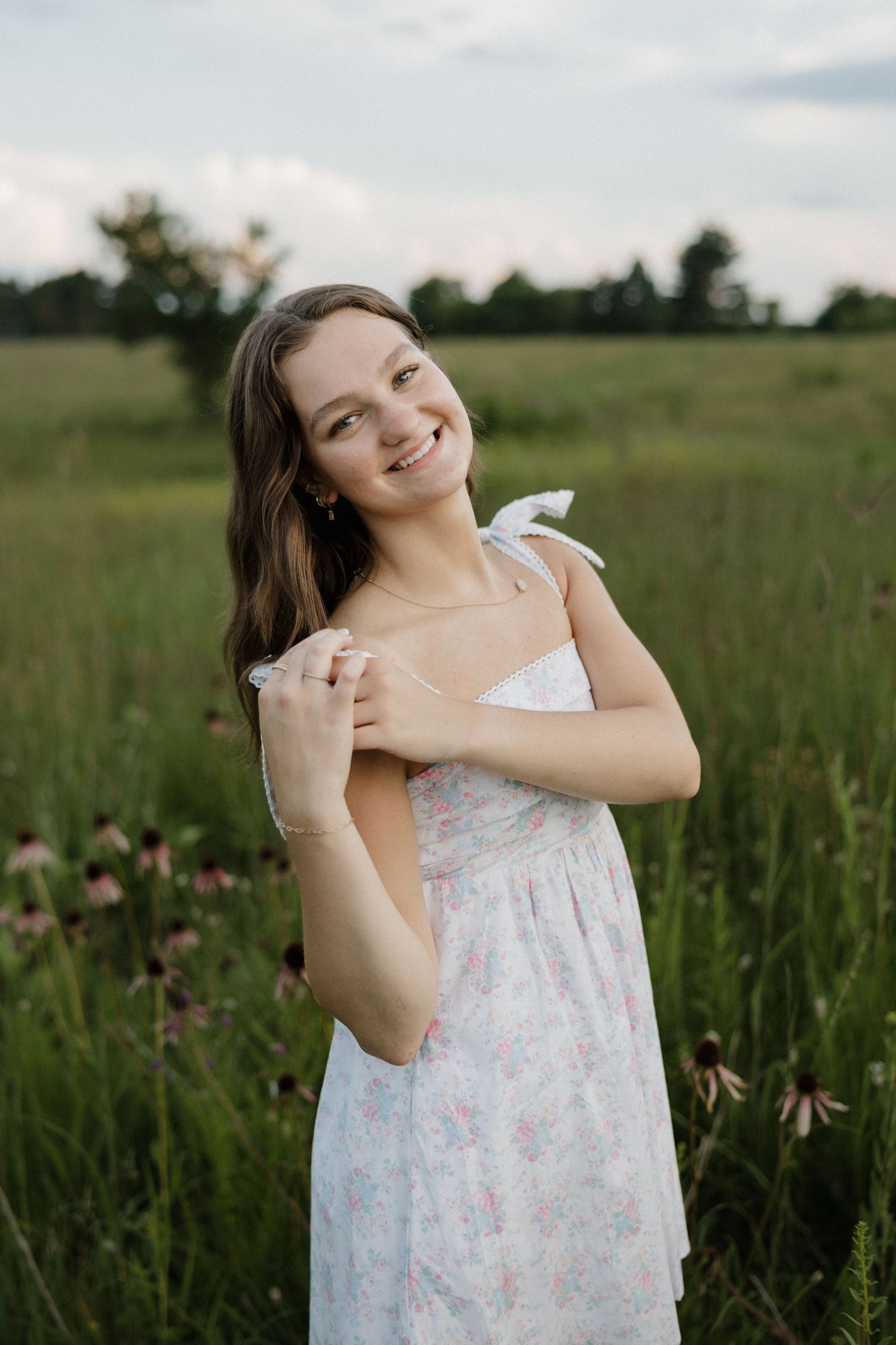 Smiling young woman in a white floral dress standing in a wildflower meadow at golden hour.